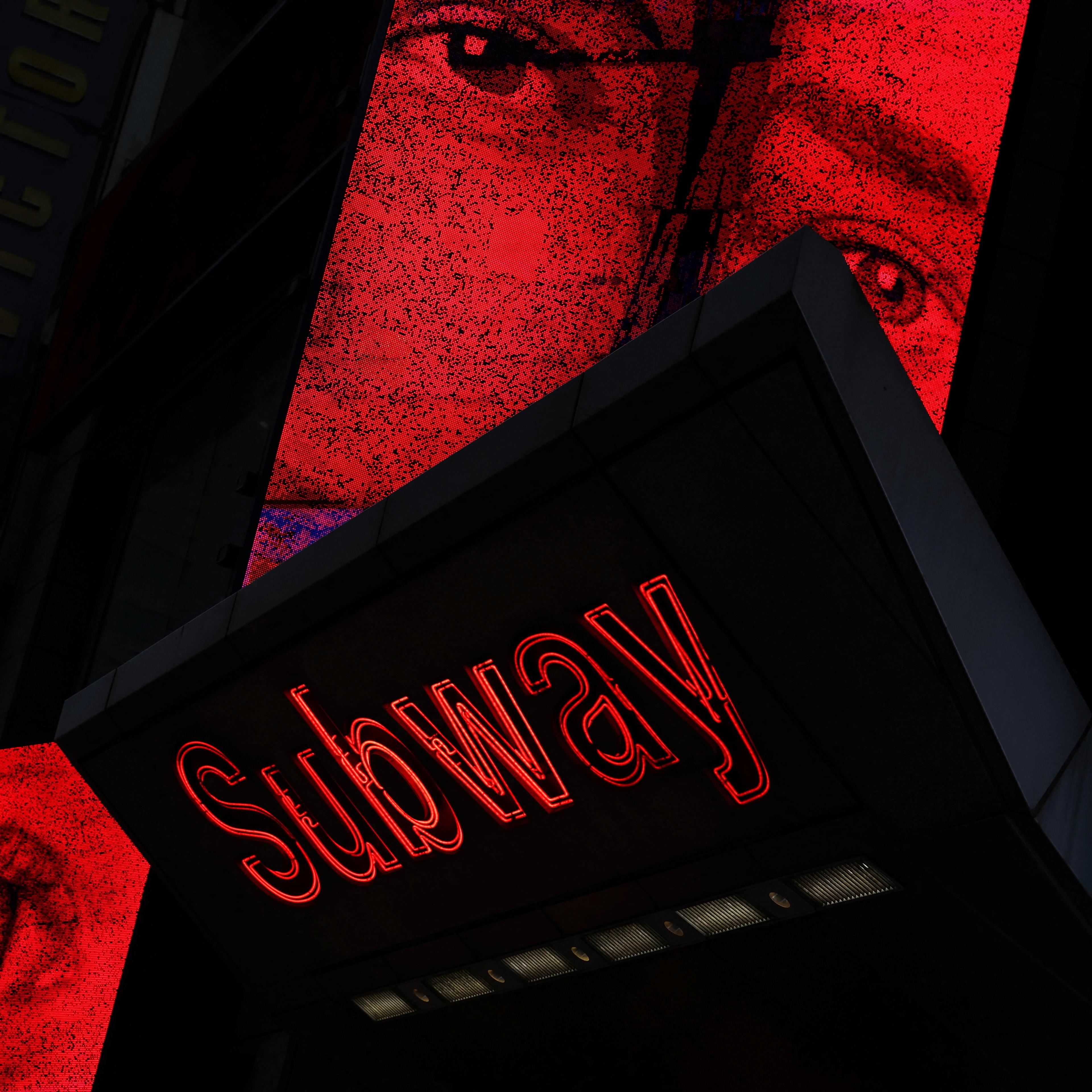 A subway sign with red neon text and large images of eyes on illuminated red panels above.