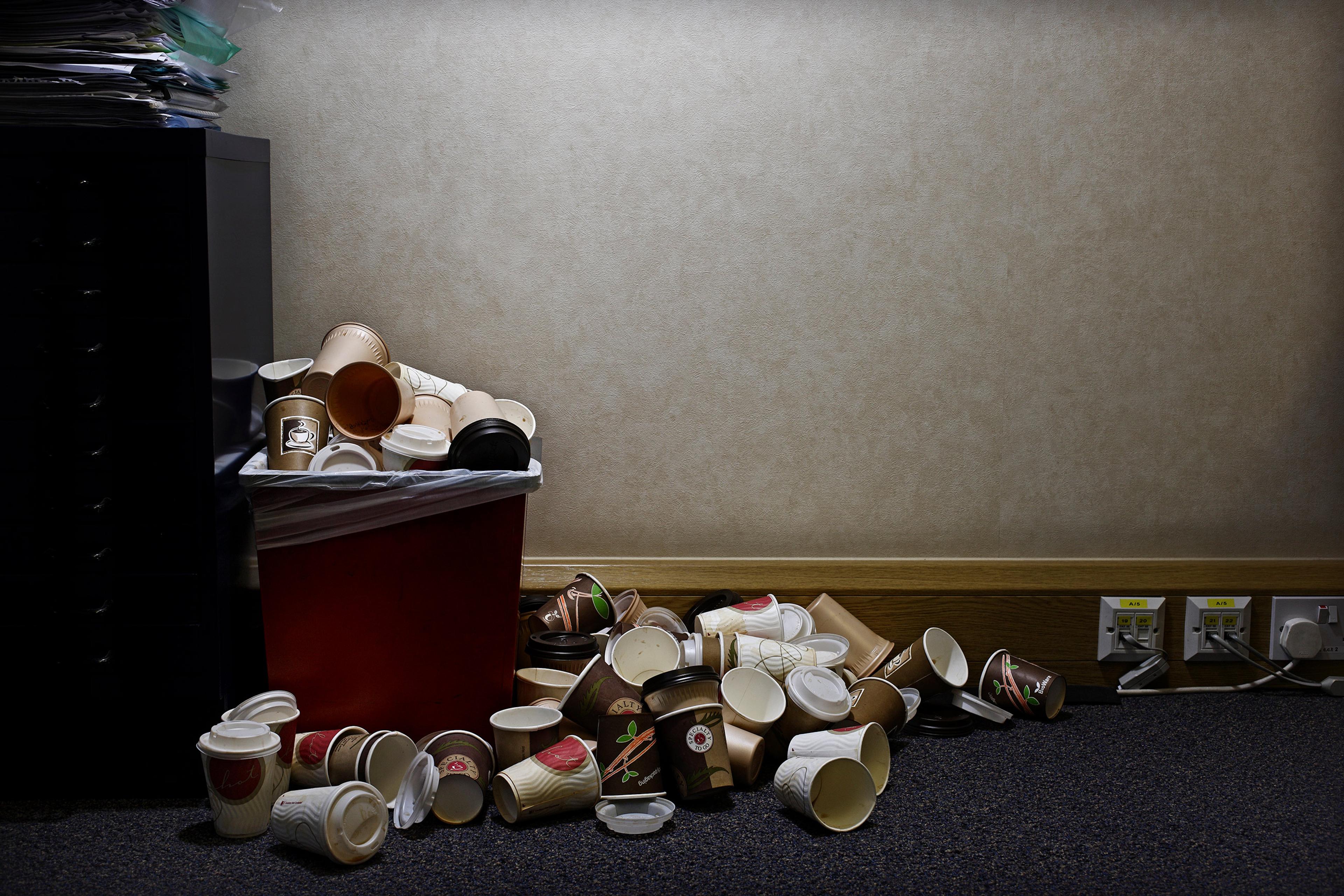 A red bin overflowing with paper coffee cups next to a wall with powerpoints in an office setting.