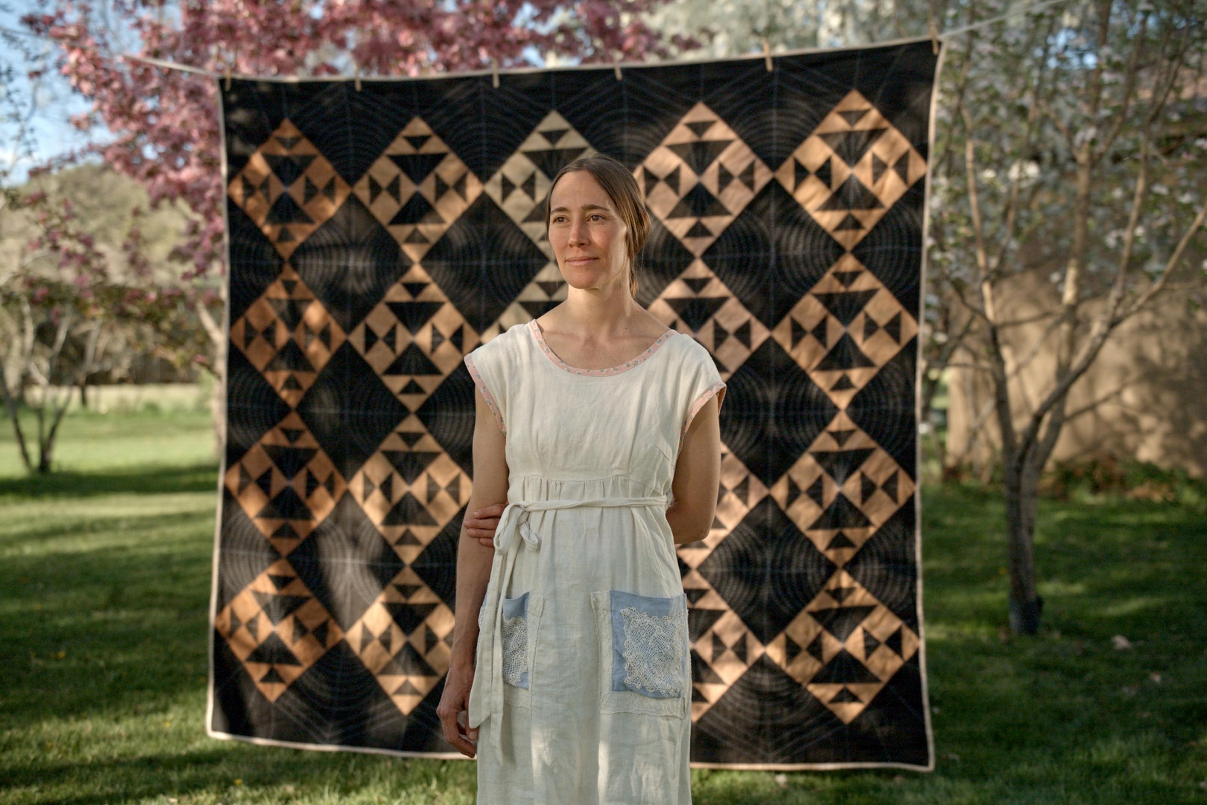 Woman in a white dress standing in front of a geometric quilt in a garden setting with trees in the background.