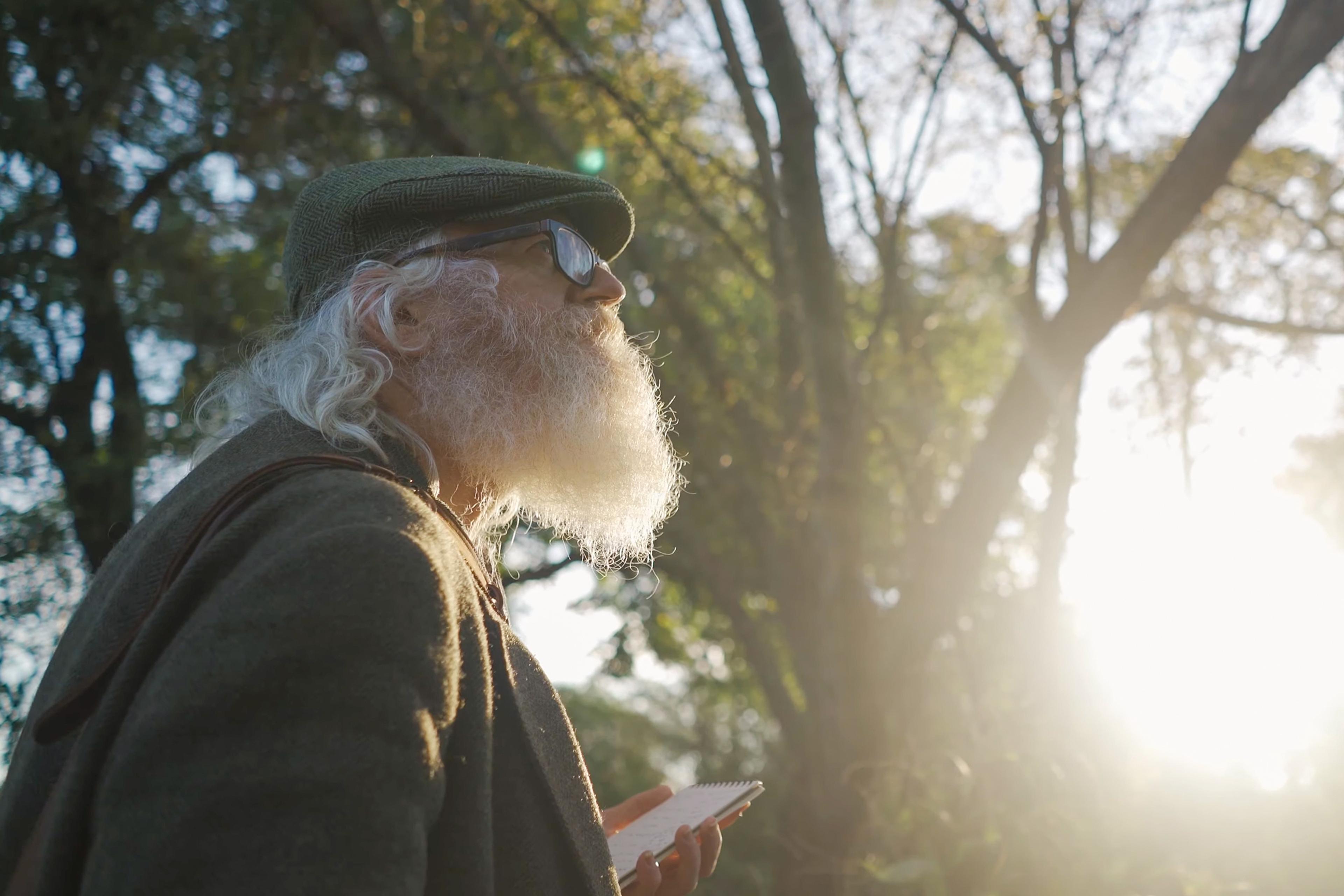 Photo of an elderly man with a beard wearing a hat and glasses, holding a notebook in a sunlit forest.