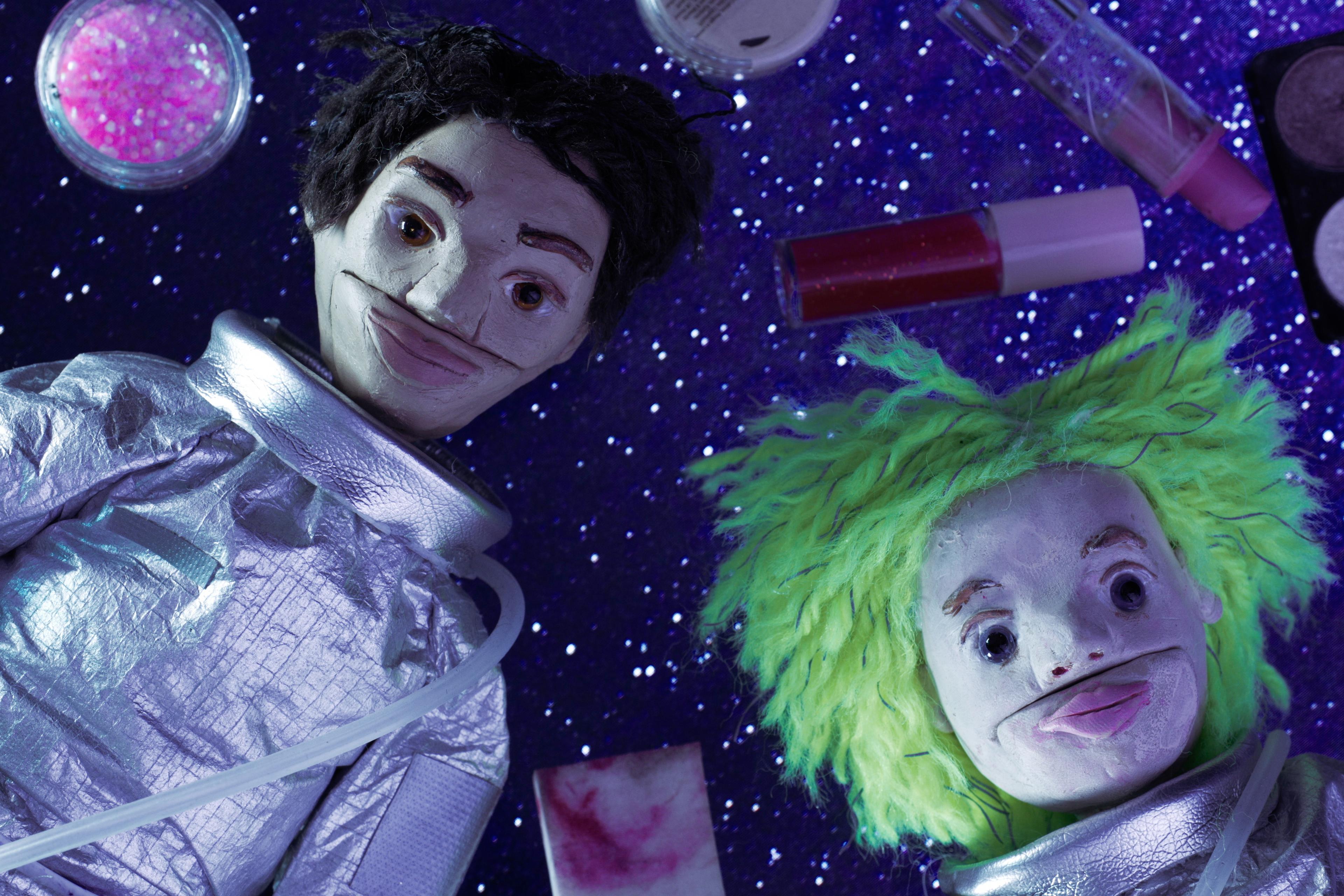 Two puppets in silver spacesuits lie against a starry backdrop, surrounded by makeup items. One puppet has short black hair, the other has bright green hair.