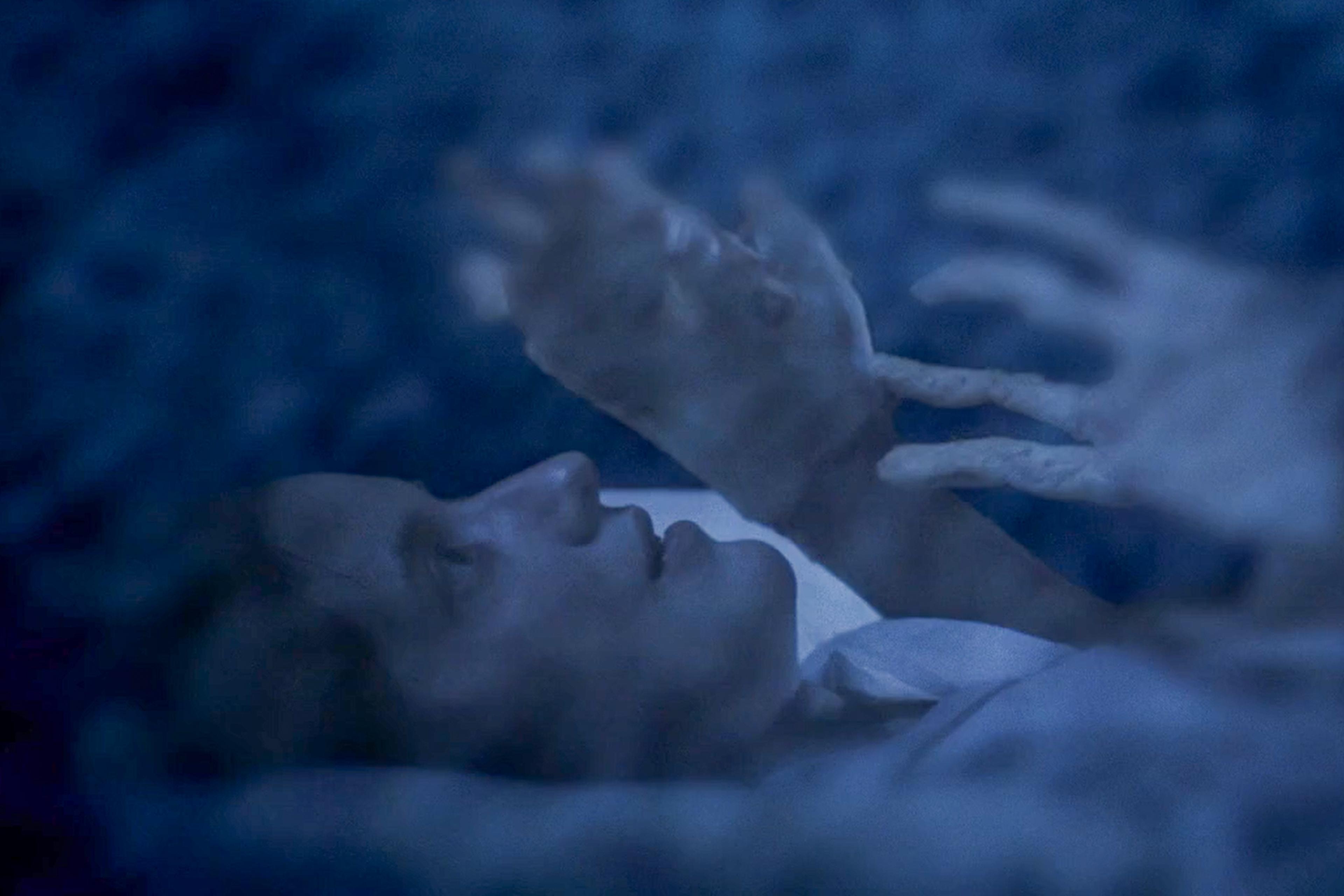 Ethereal photo of a person lying down under a blue haze with ghostly hands reaching towards them.