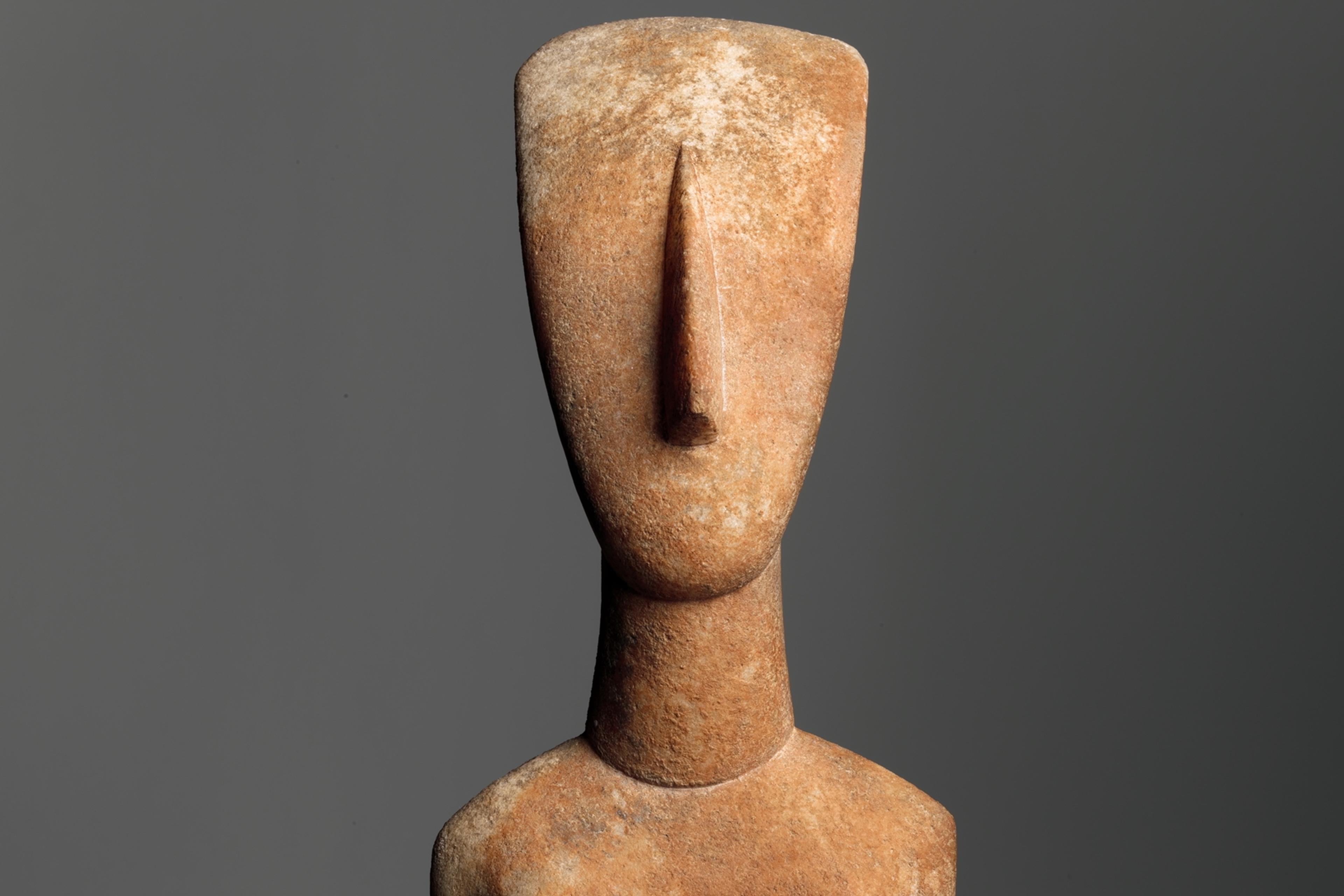 Abstract stone sculpture of a human figure with folded arms against a grey background.