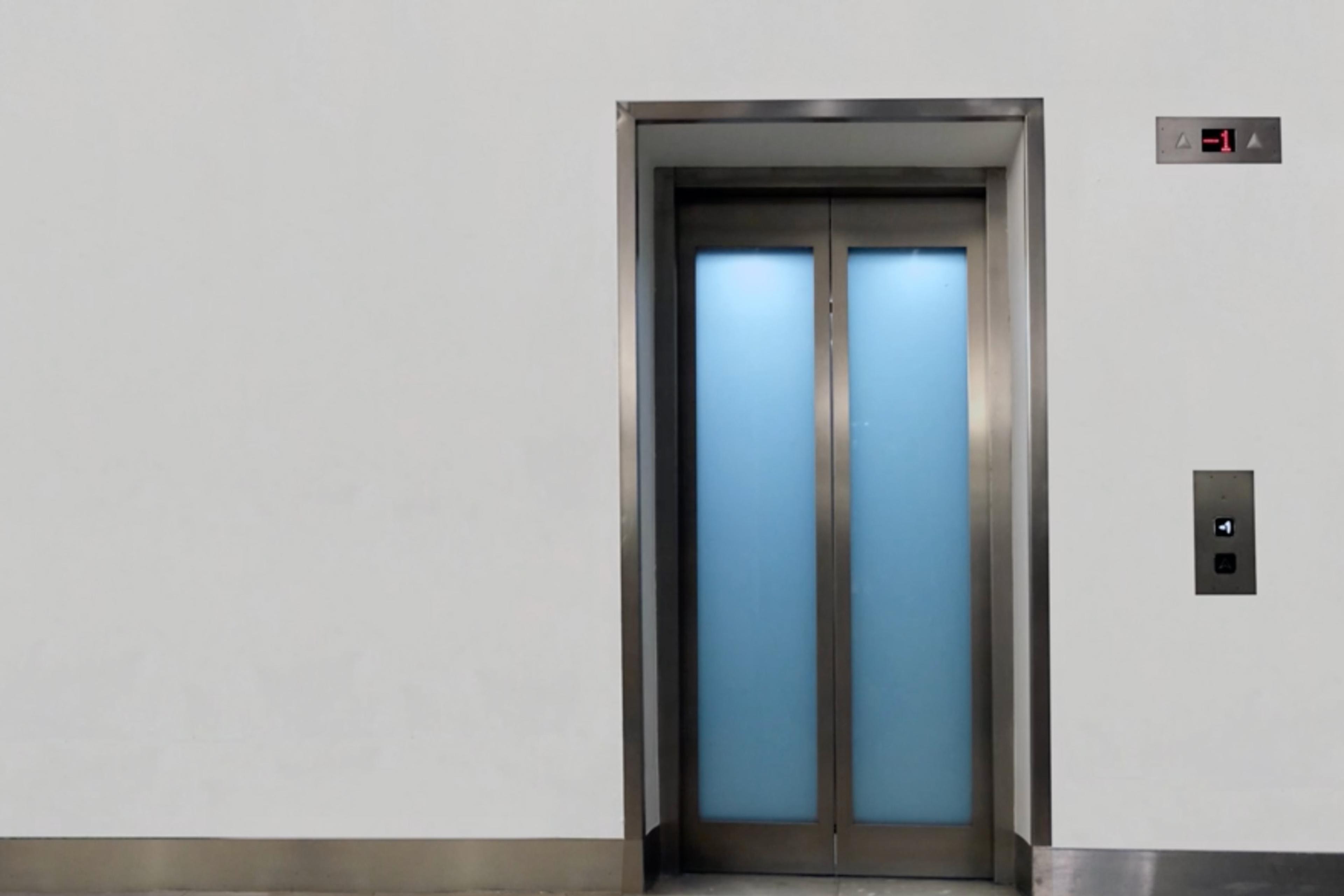 A closed lift with stainless steel doors and a blue panel display showing floor -1 above the button against a white wall.