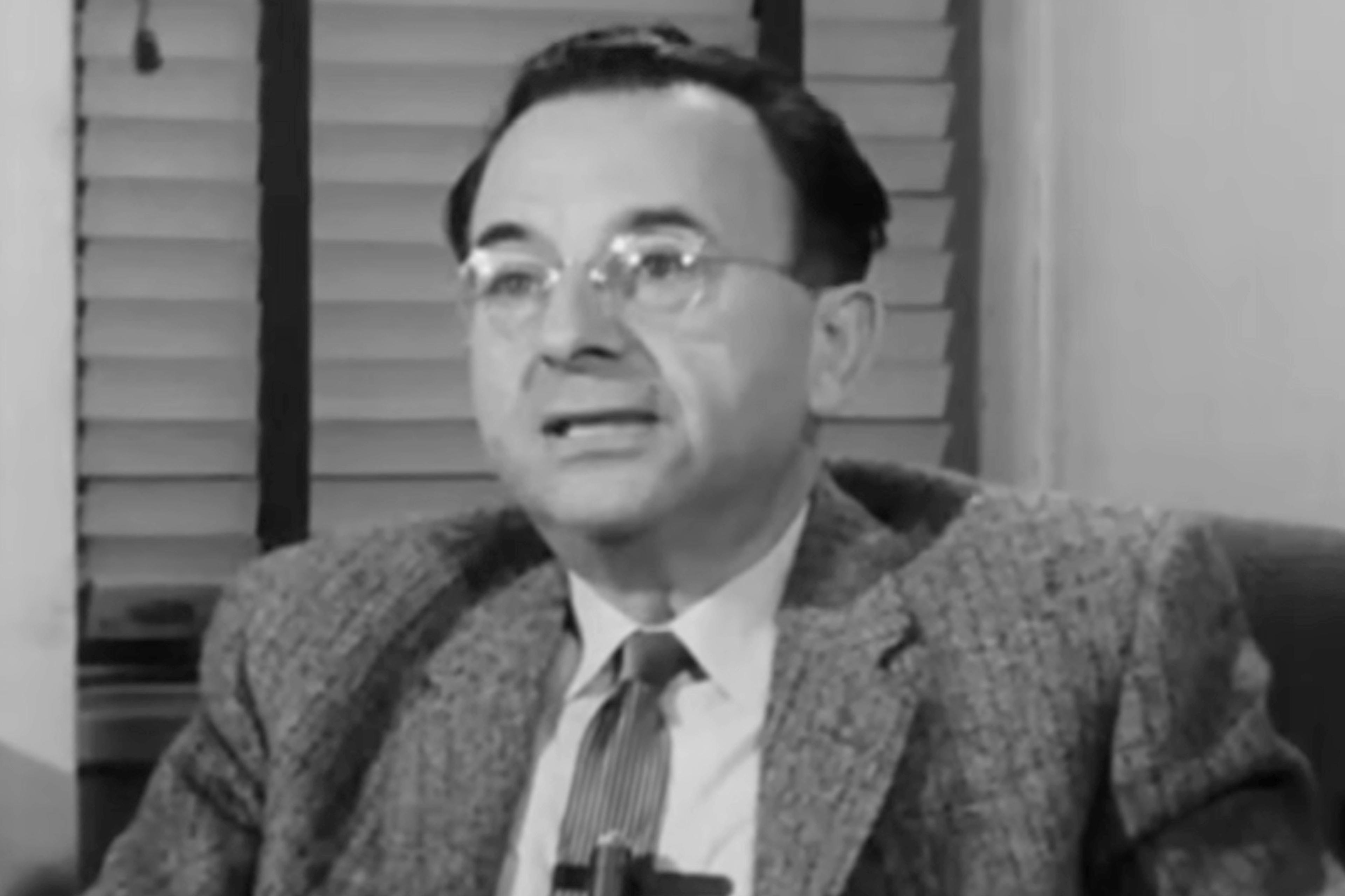 Black and white photo of a man in glasses and a suit speaking, seated indoors with window blinds in the background.