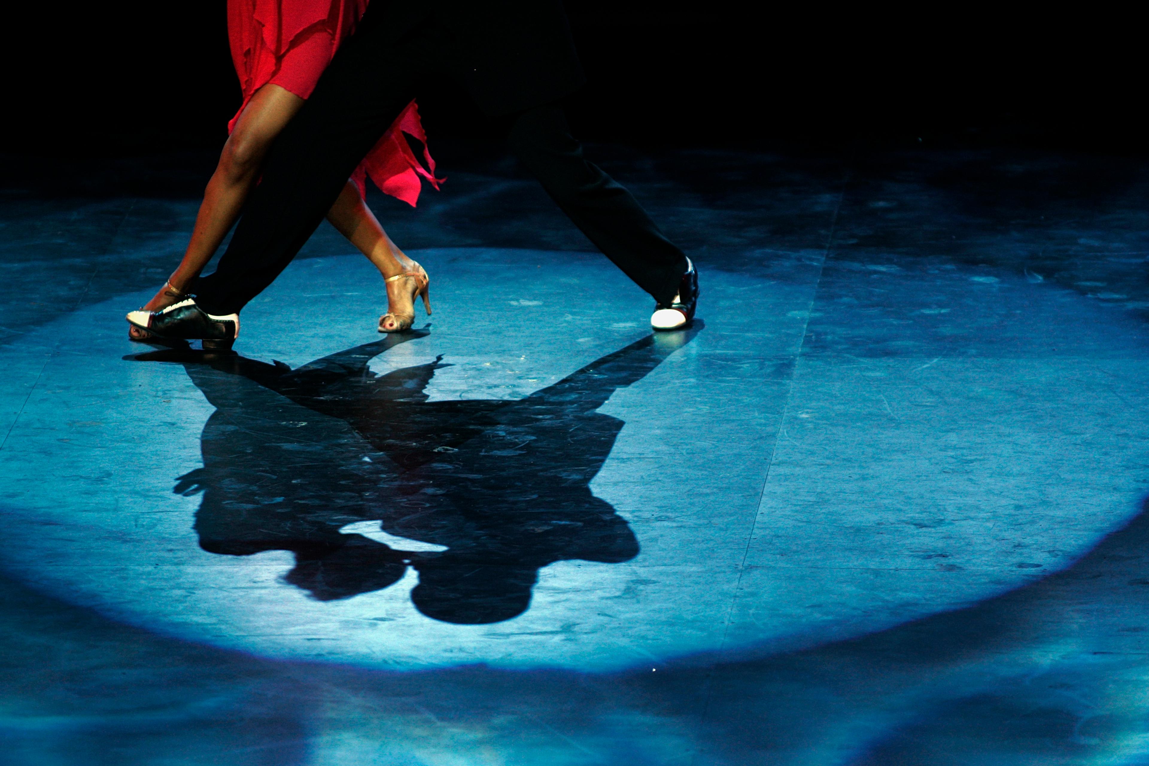 A couple dancing on stage, the photo focusing on their feet and shadows, with dramatic lighting and a blue spotlight.