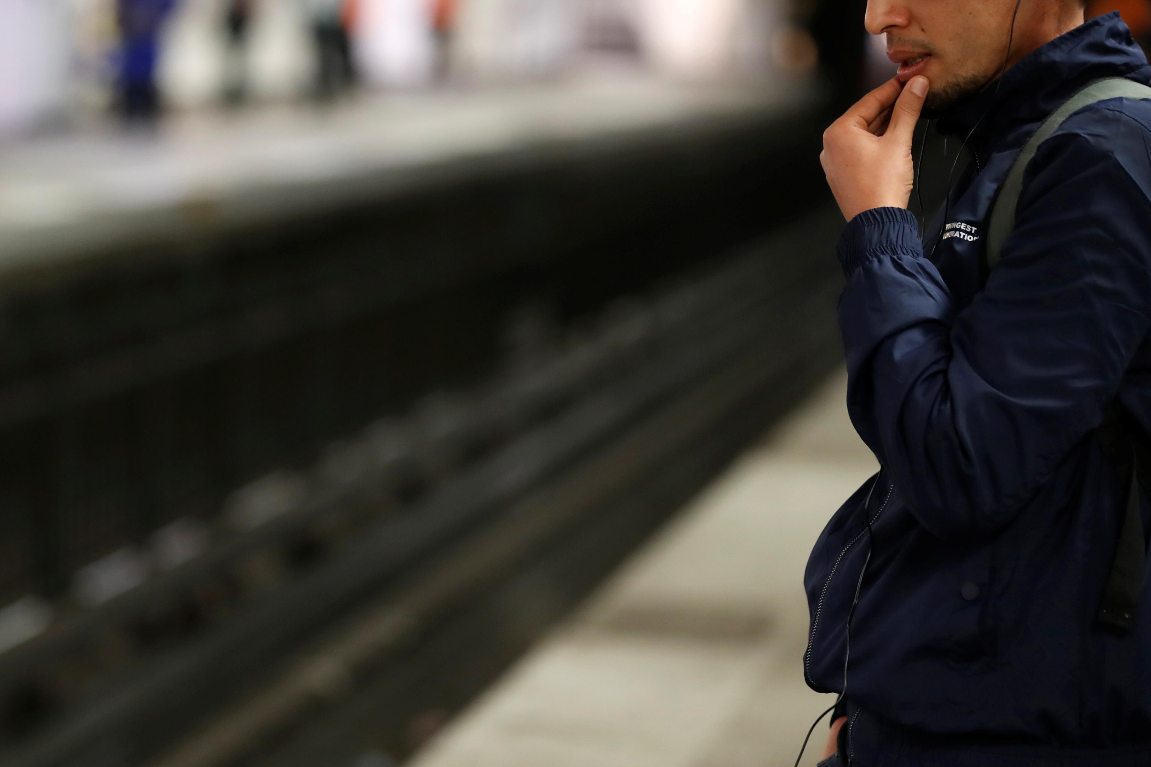 A person in a blue jacket standing thoughtfully on a train platform next to the tracks.