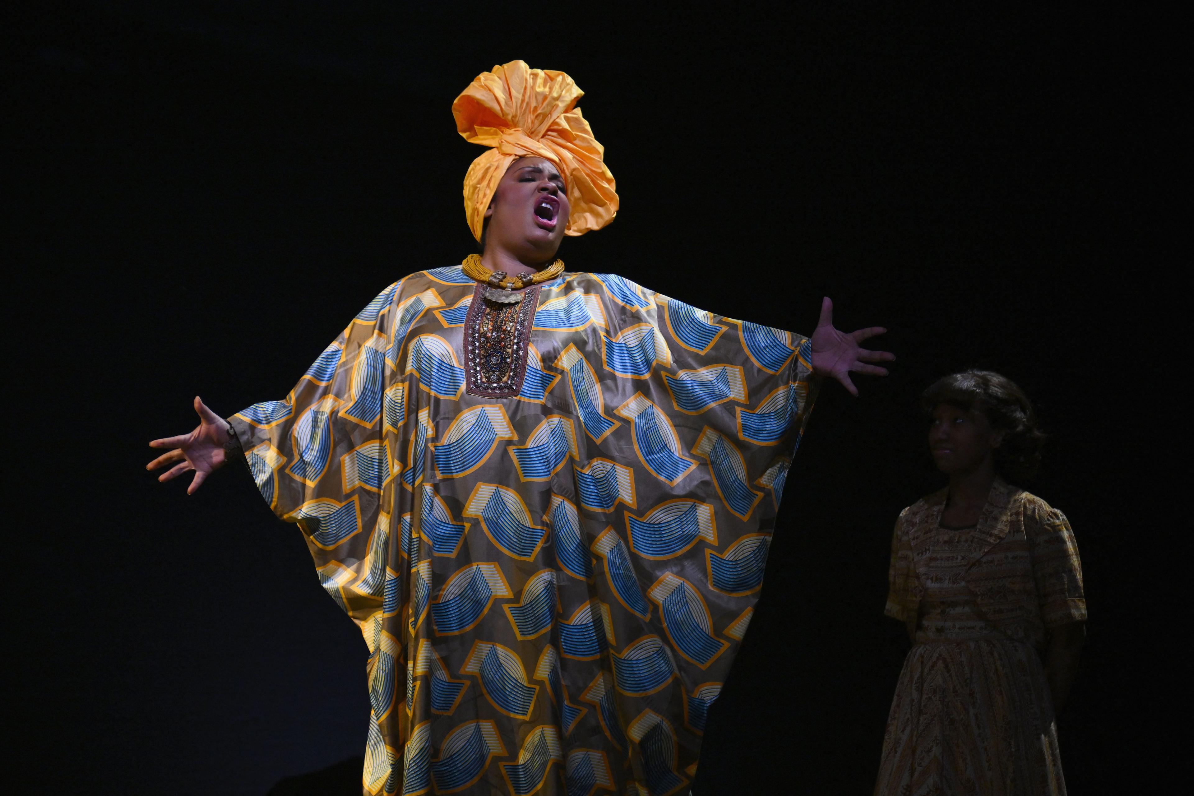 Photo of a person in vibrant traditional attire with an orange headwrap singing on stage, another in shadow to the side.