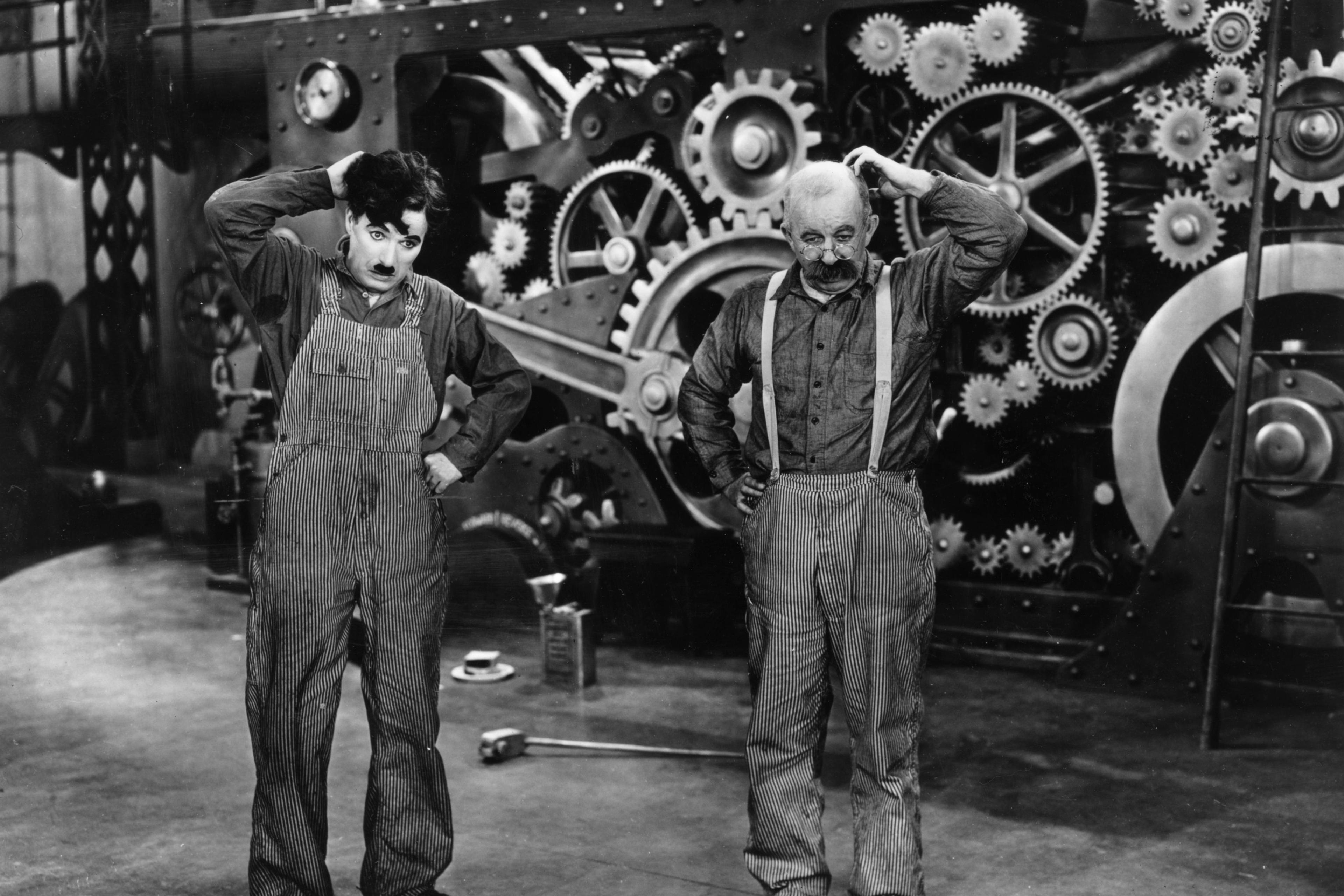 Black and white photo of two men in overalls standing before large industrial gears, each scratching his head.