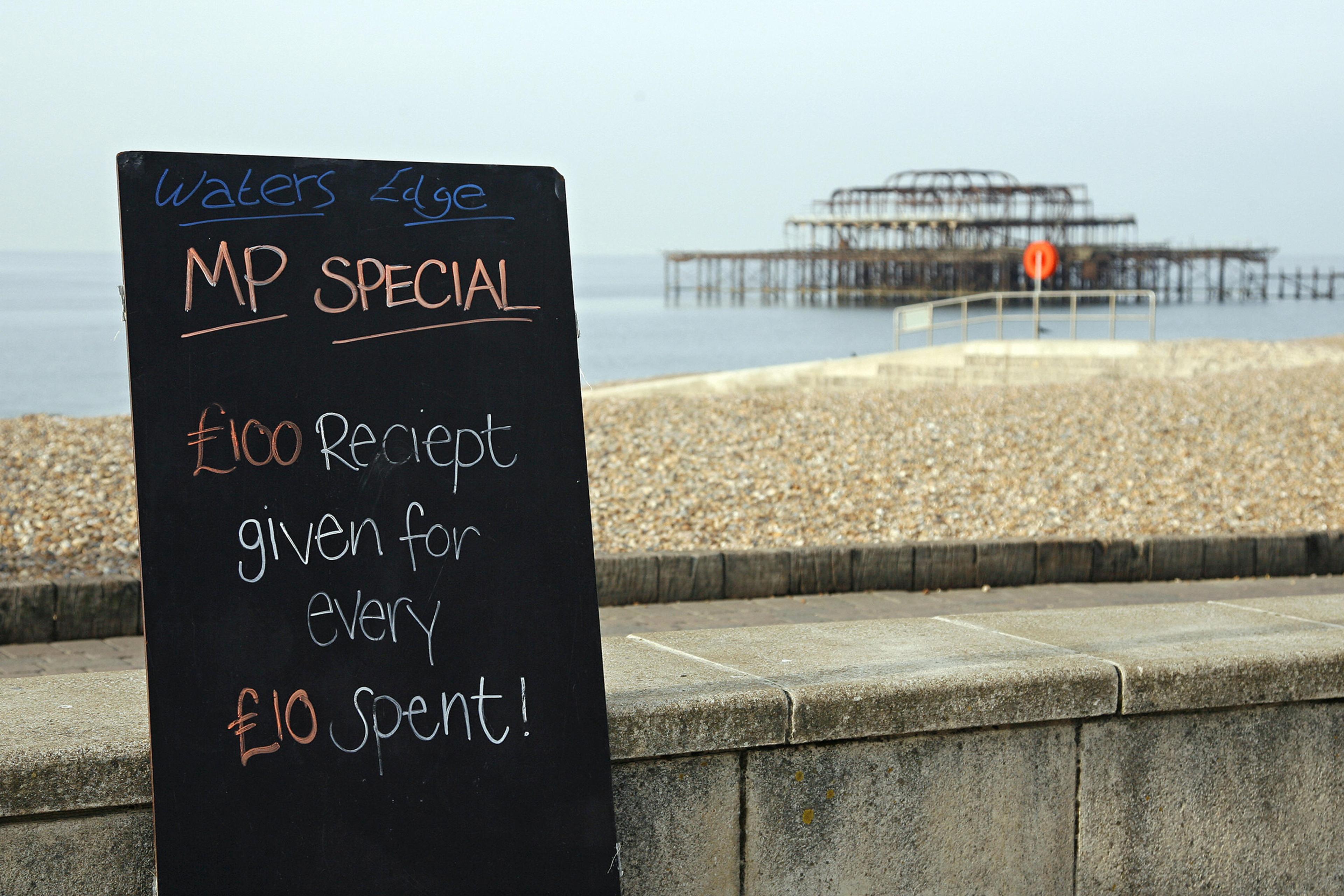 Photo of a beachside blackboard offering a humorous deal near a weathered pier, stating “£100 receipt given for every £10 spent.”