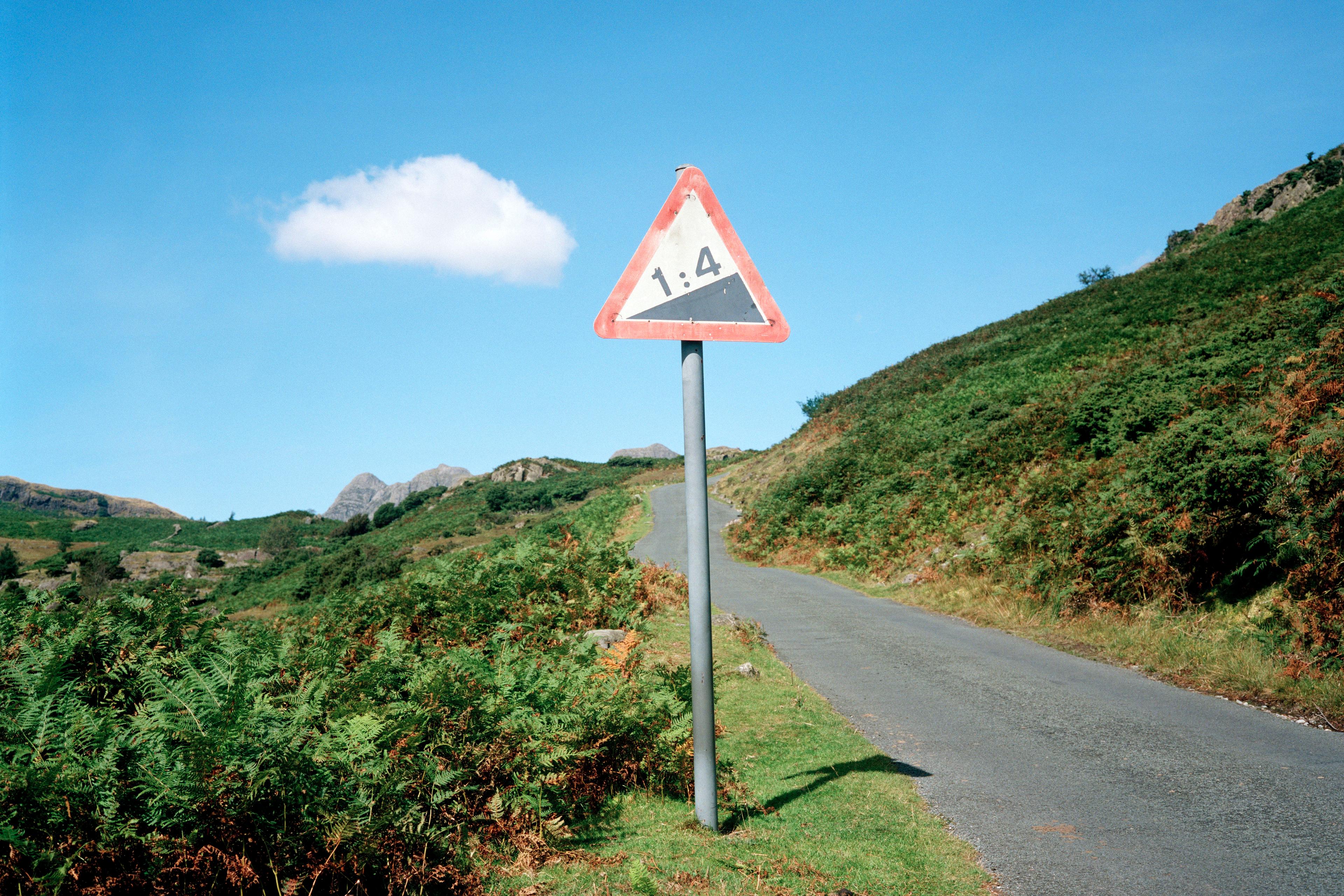 Photo of a rural road with a steep hill warning sign marked 1:4 surrounded by green landscape under a blue sky.