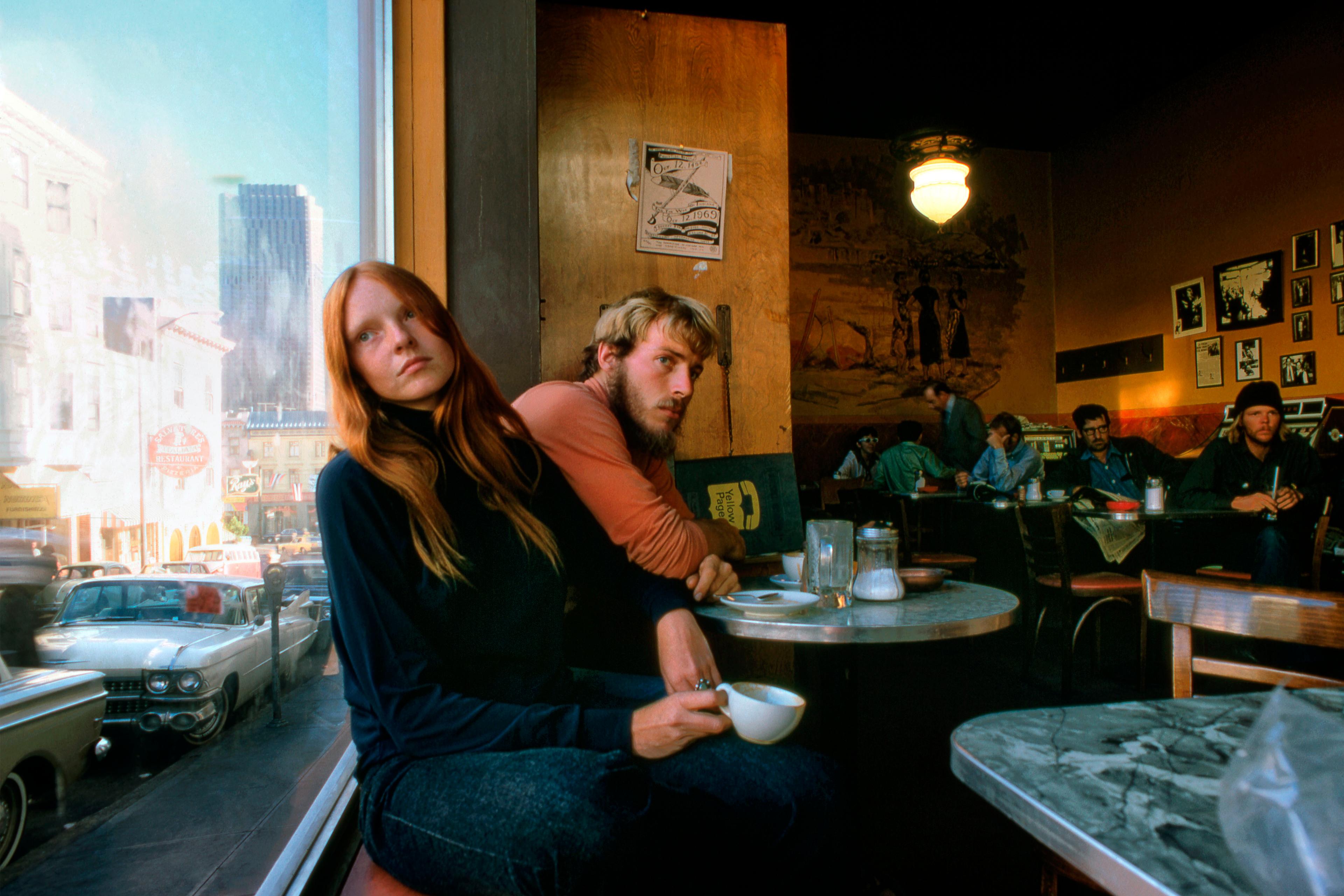 Photo of two people sitting in a cafe by a window, with street view and classic cars outside. Warm lighting inside.