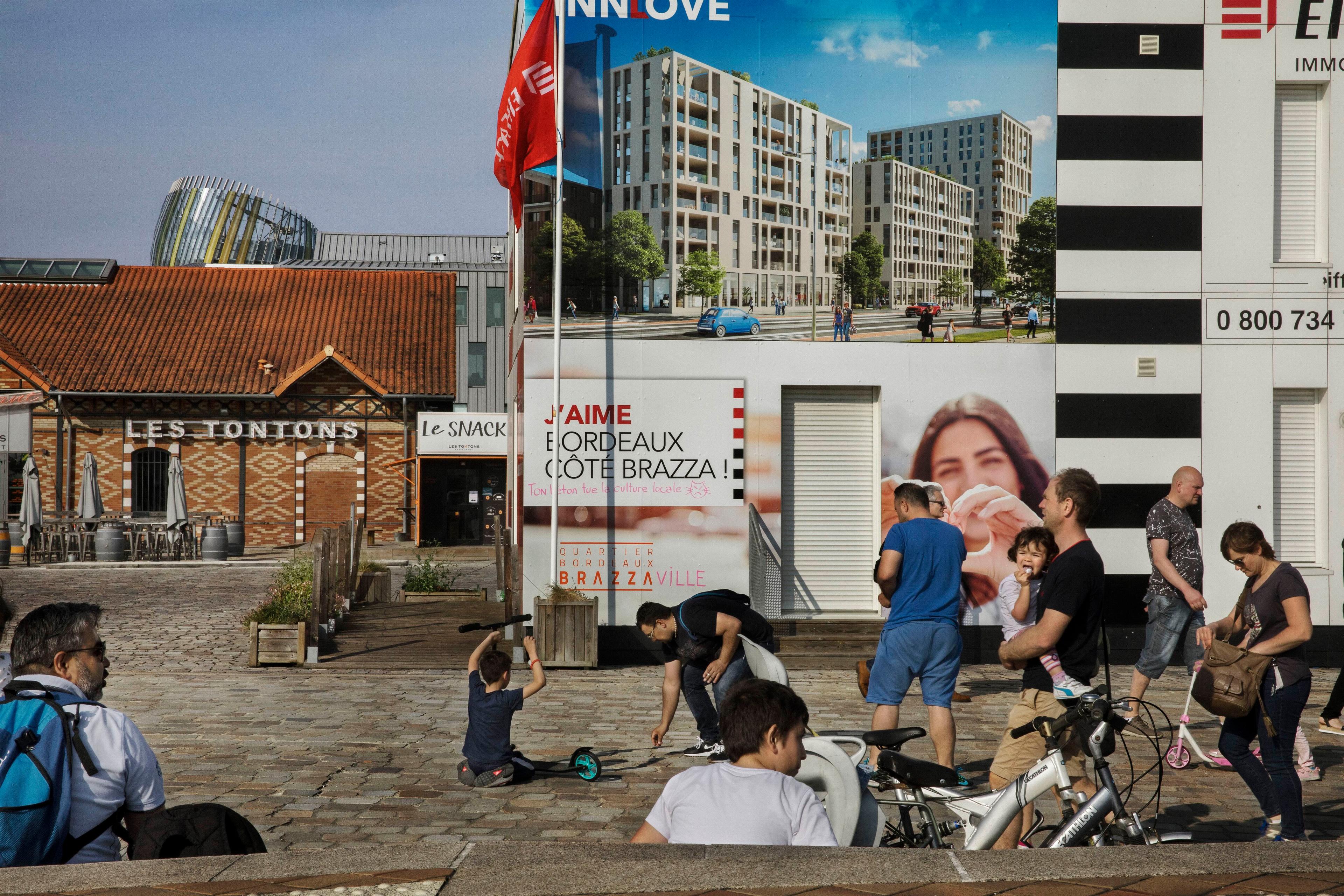 Photo of people in a sunny square with bikes and scooters, near Les Tontons and a large building advertisement in Bordeaux.