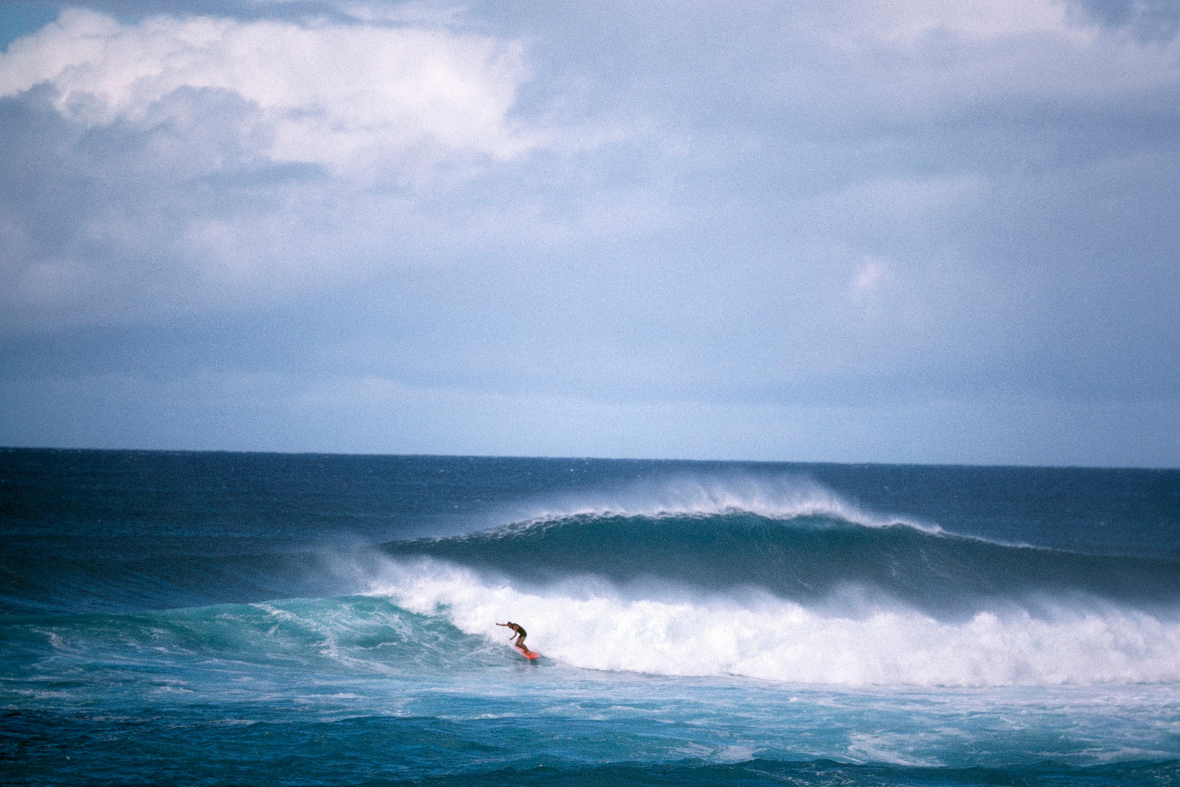 A surfer riding a wave under a cloudy sky, with another large wave shown behind.