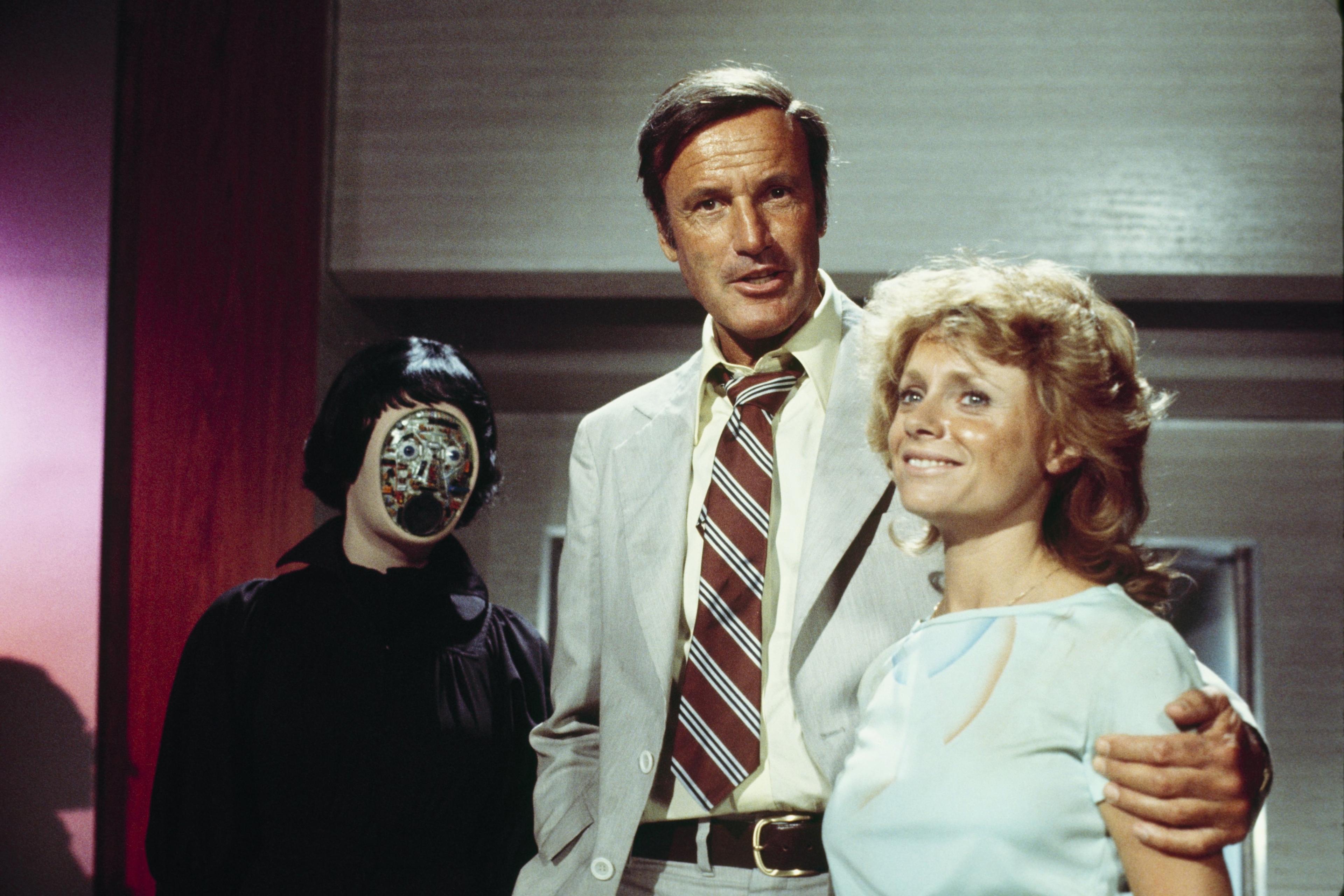Photo of a man in a suit with two people, one with a mechanical face, indoors.
