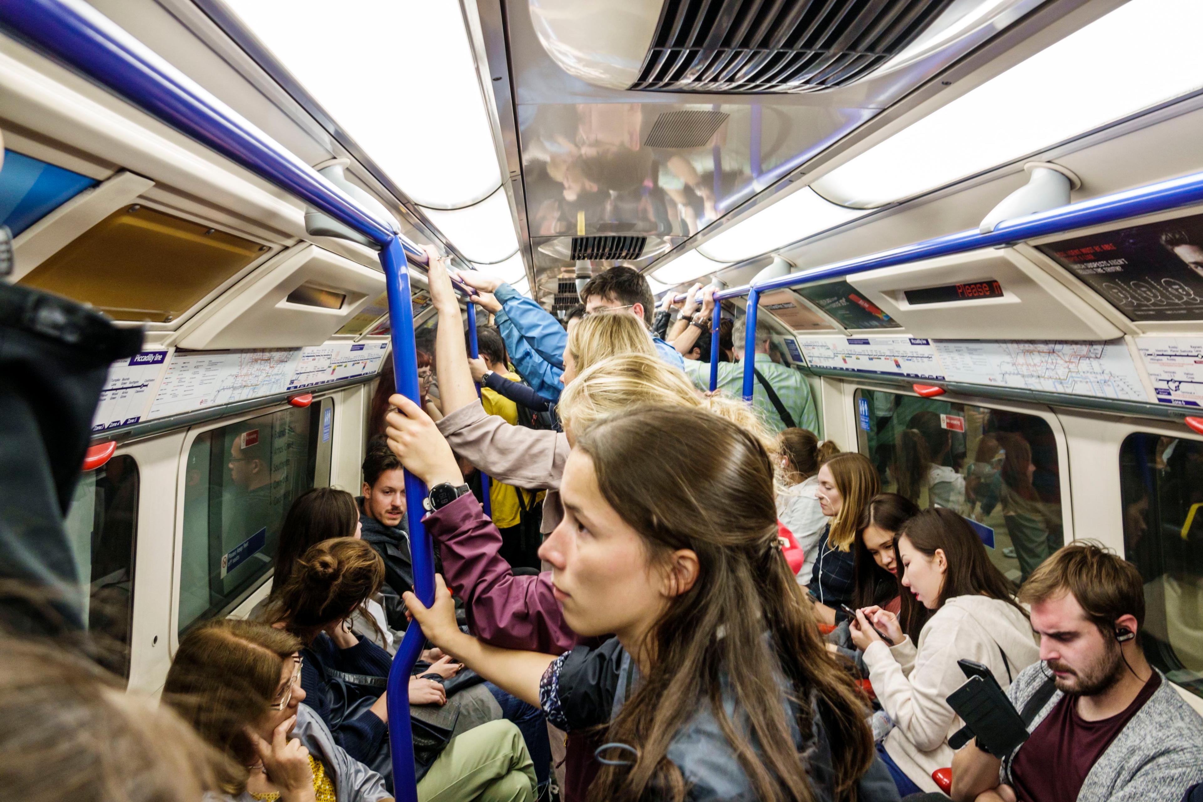 Photo of a crowded London Underground train with passengers standing and sitting, holding onto poles and looking at phones.
