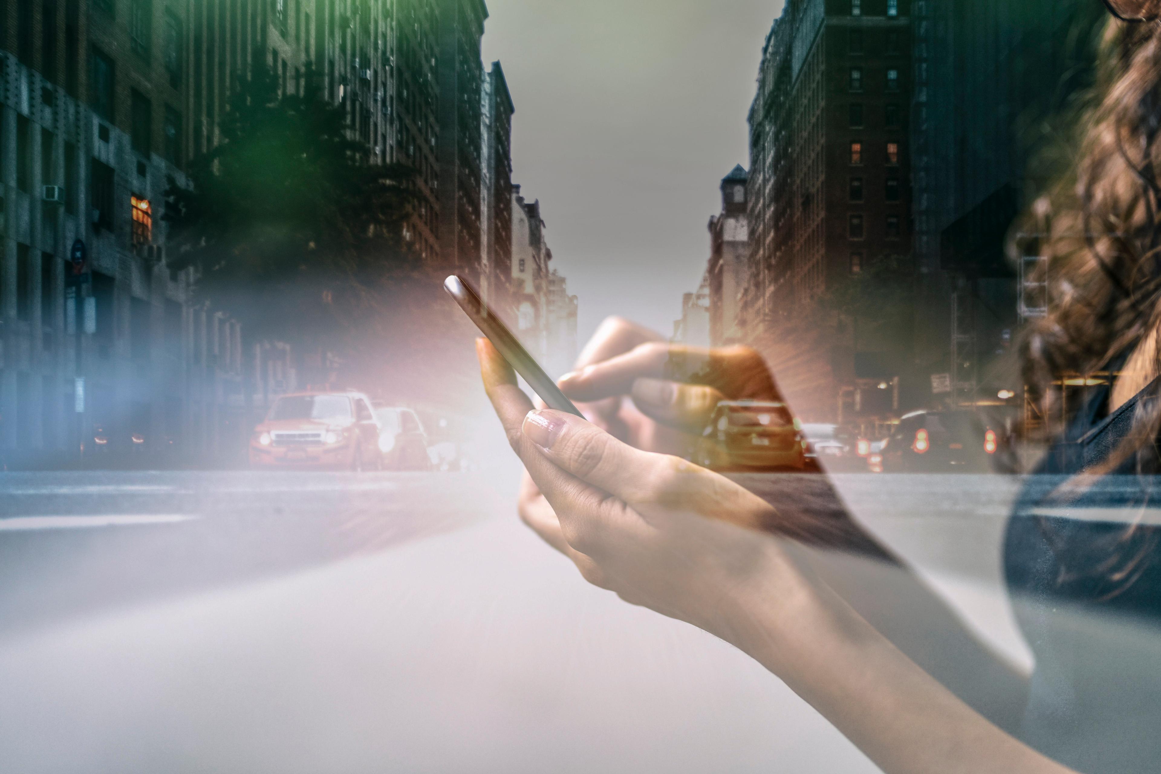 Photo of a hand using a smartphone with a city street in the background, creating a double exposure effect.