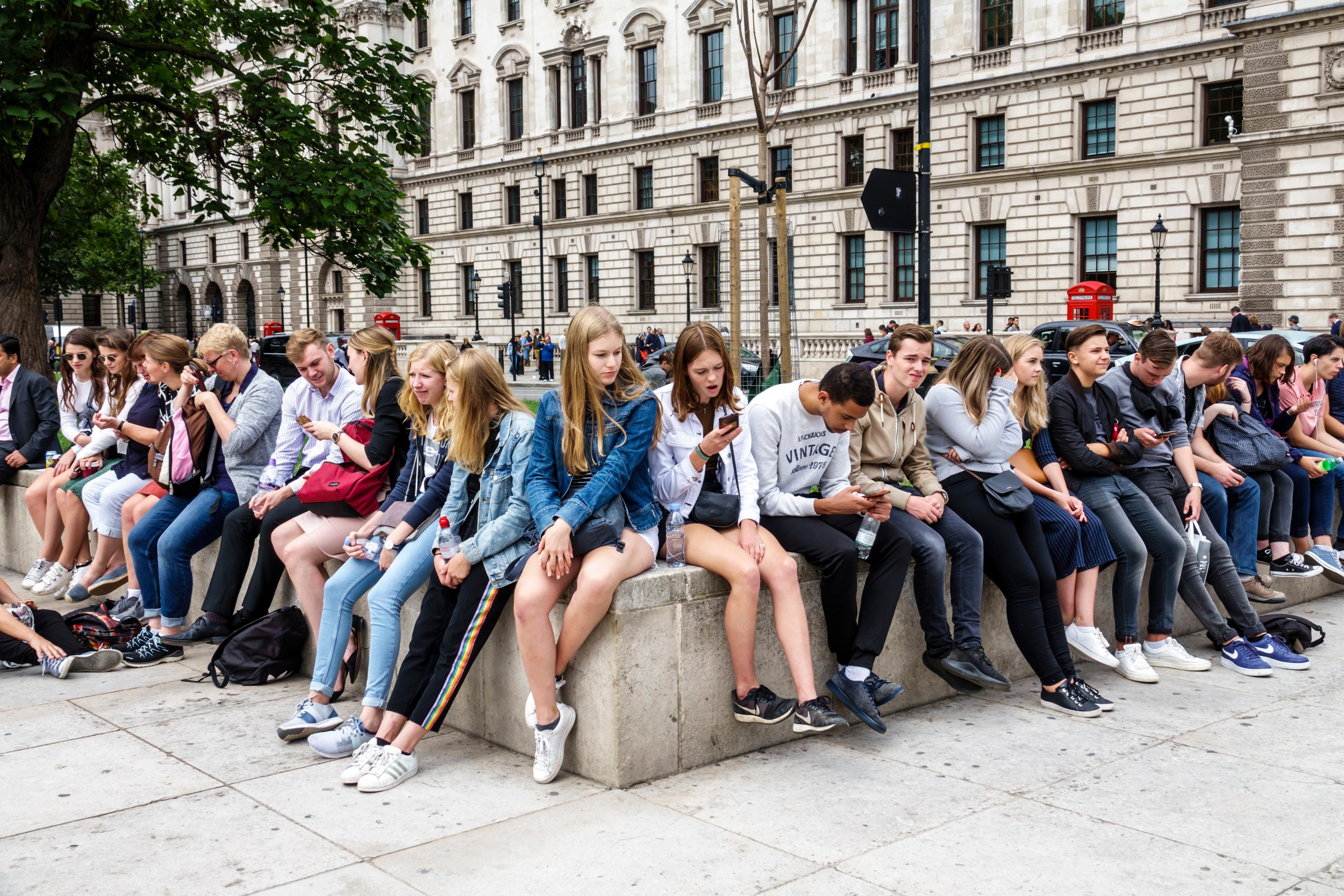 Photo of young people sitting on a stone ledge outside a historic building, some using phones, with trees and red phone boxes.