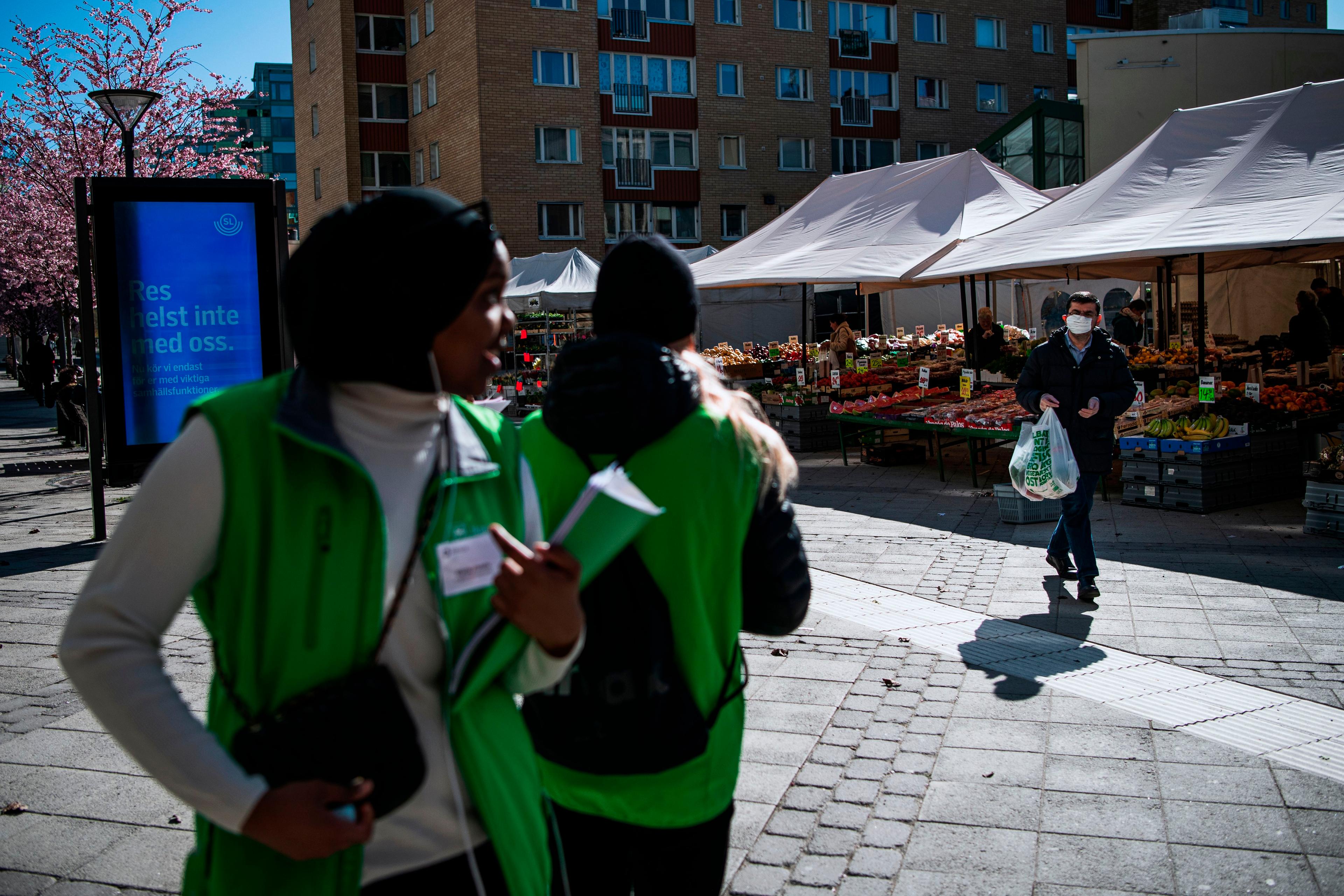 Photo of an outdoor market with fruit stalls, a masked man carrying bags and two people in green vests walking past.
