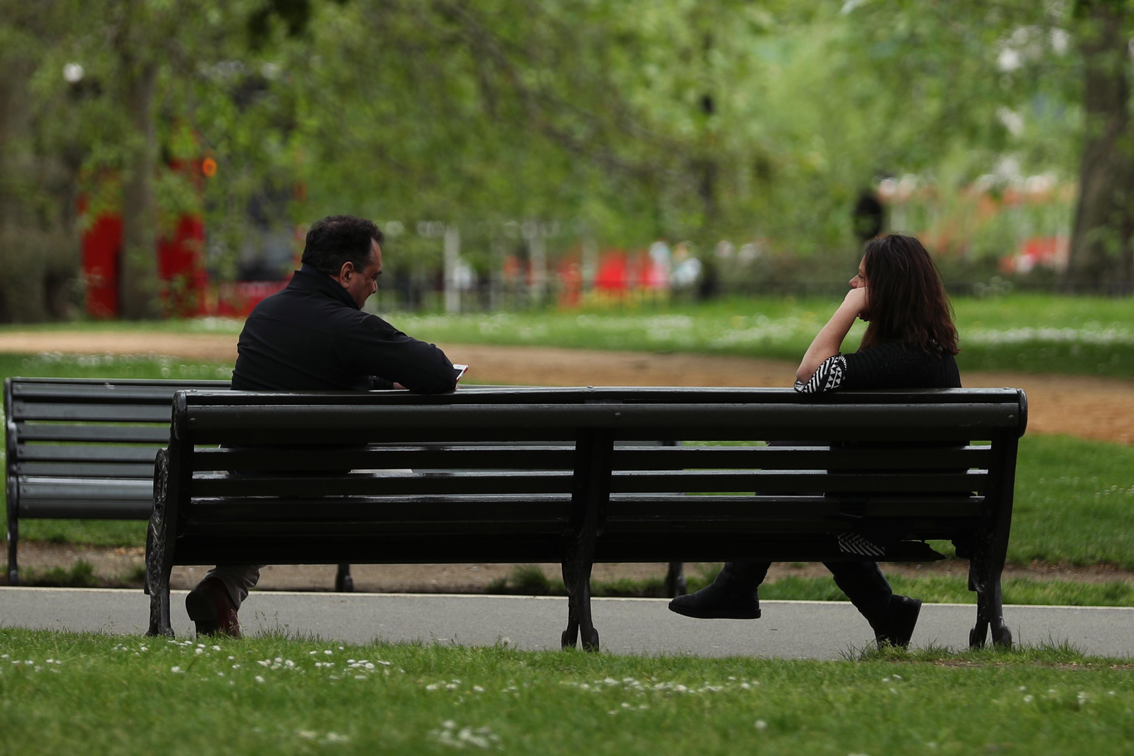 Photo of two people sitting on a park bench in a lush green setting with a pathway and trees in the background.