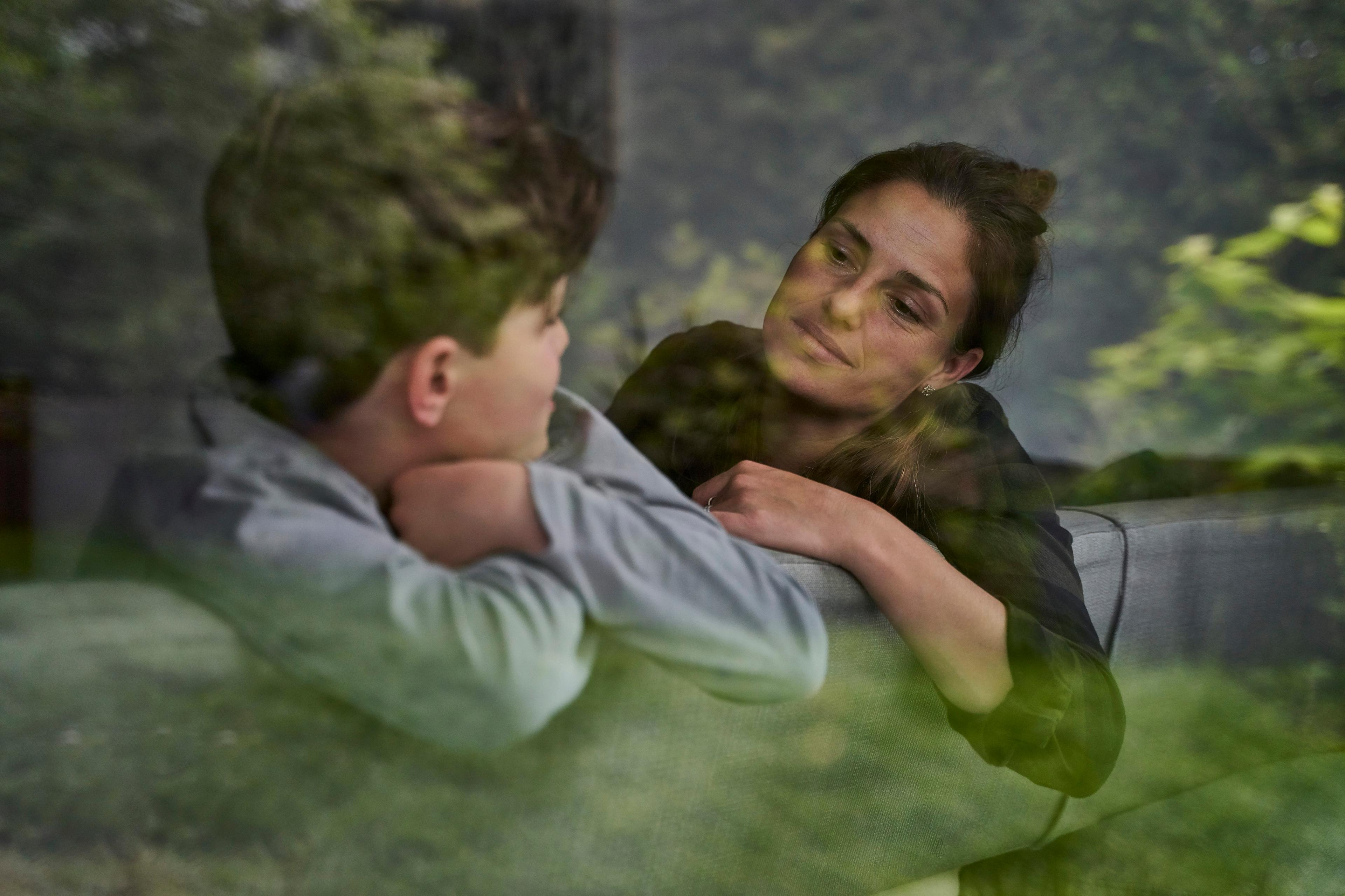 Photo of a woman and child talking, seen through glass with reflections of greenery outside.
