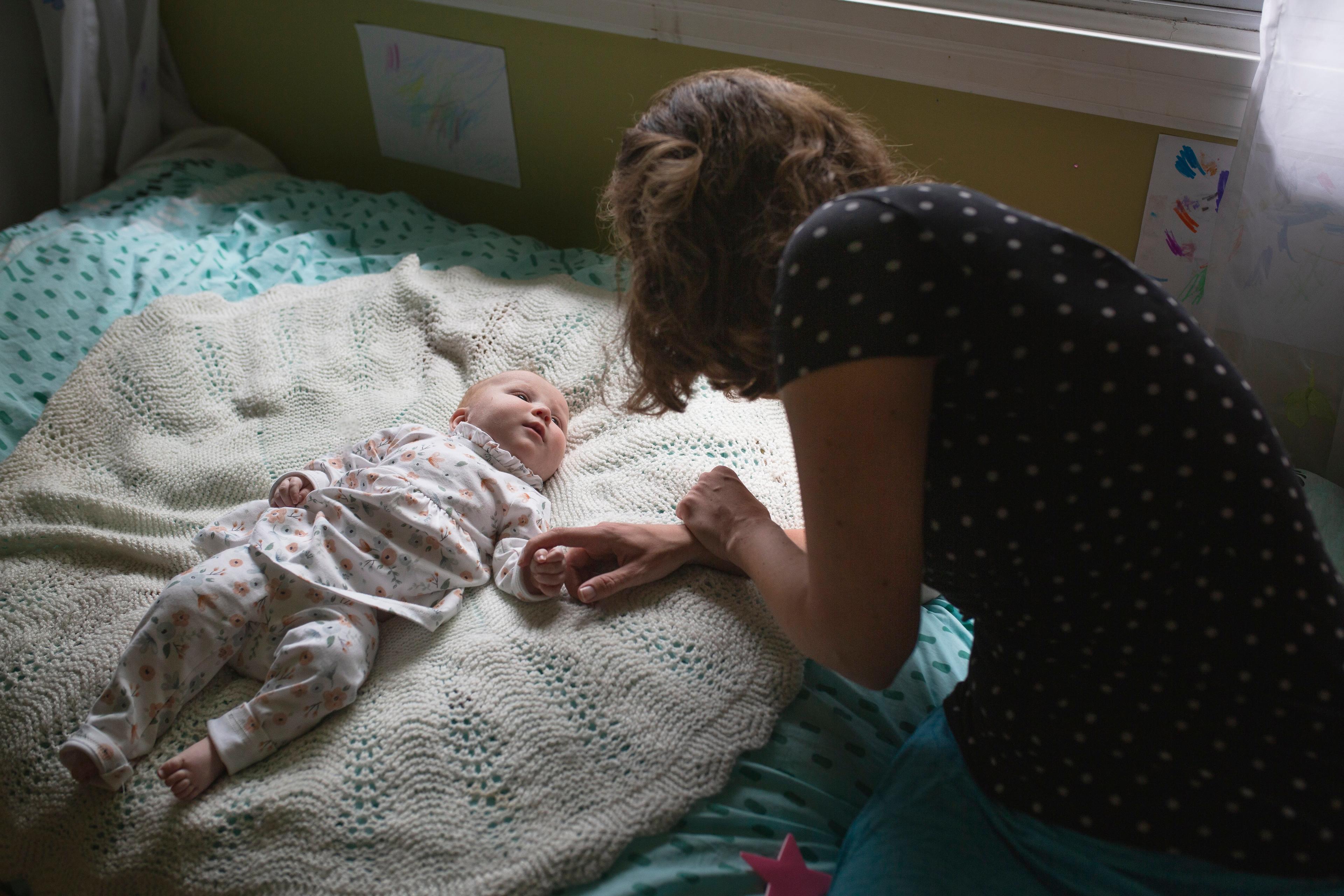 Photo of a woman leaning over a baby lying on a bed, holding the baby’s hand in a softly lit room.