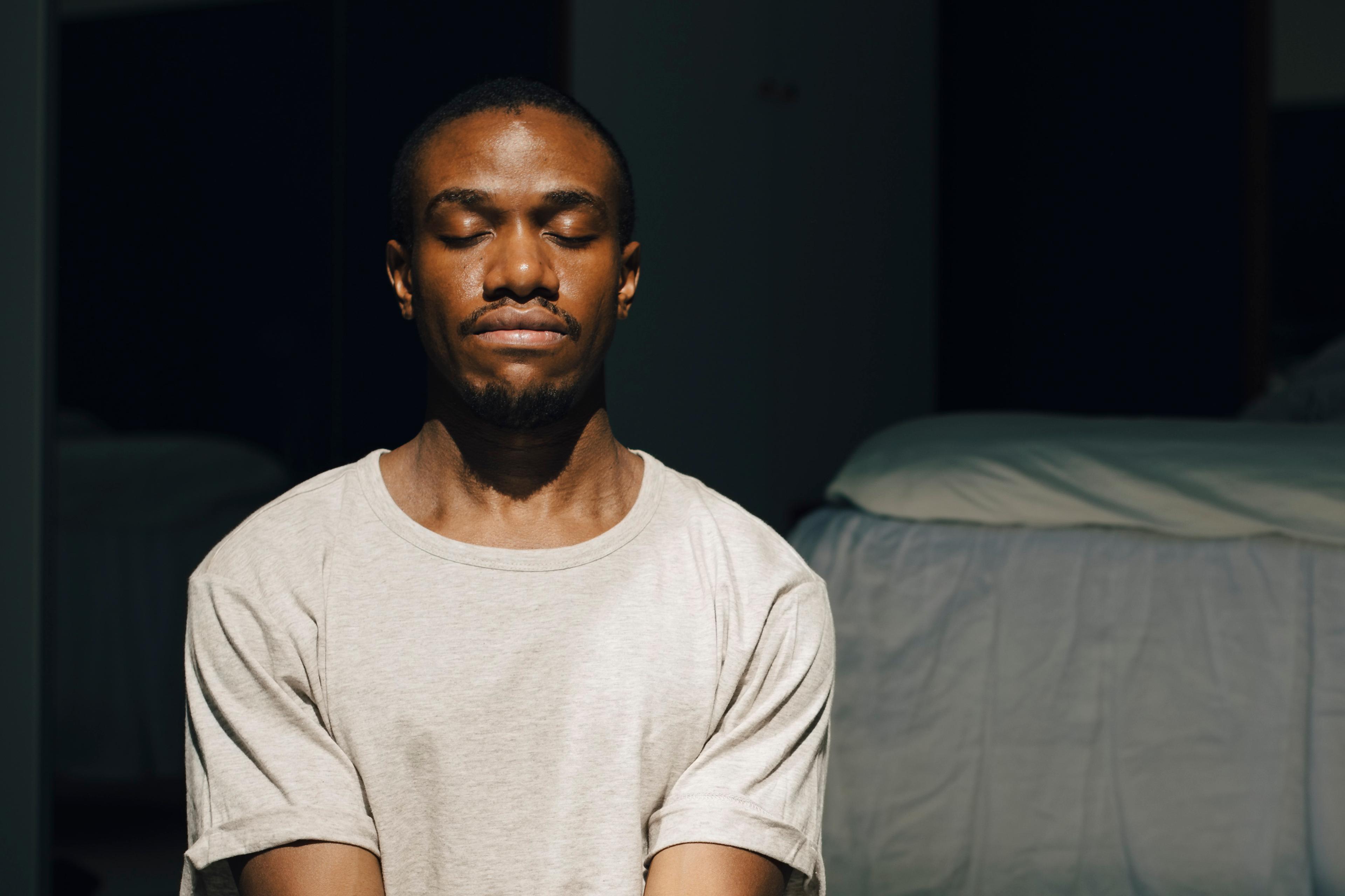 Photo of a person meditating in sunlight on the floor of a room beside a bed.