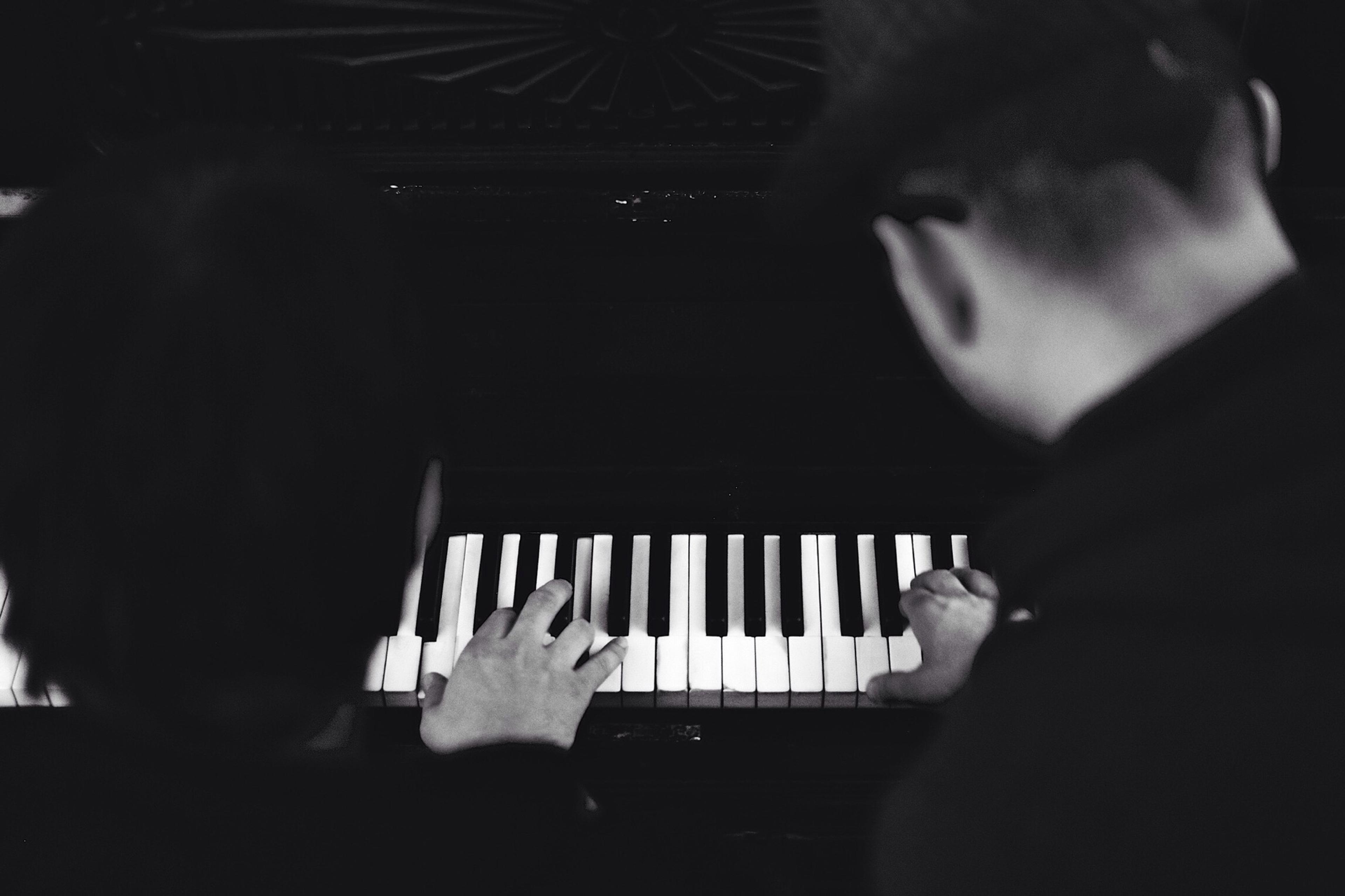 Black and white photo of two people playing a piano, focused on their hands and the keyboard from above.