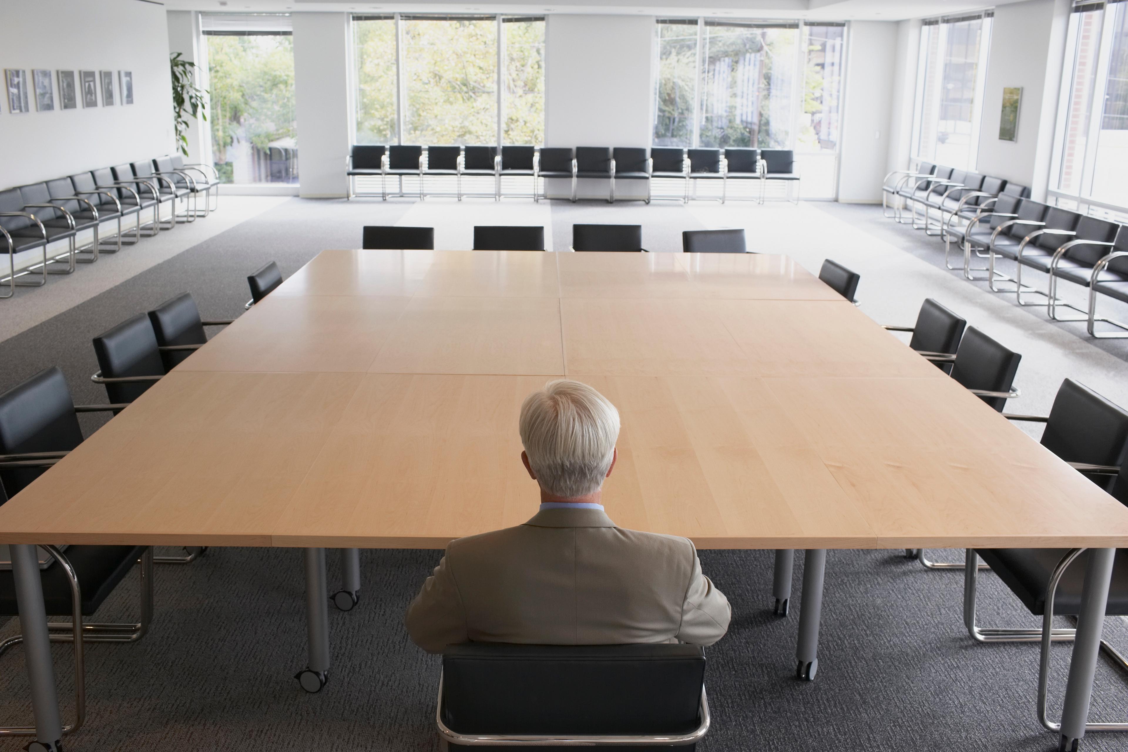 A man with white hair in a suit sitting at the head of an empty boardroom table, facing forward, back toward the camera. Dozens of empty chairs line the outer edges of the room; windows at the rear of the room show trees outside.