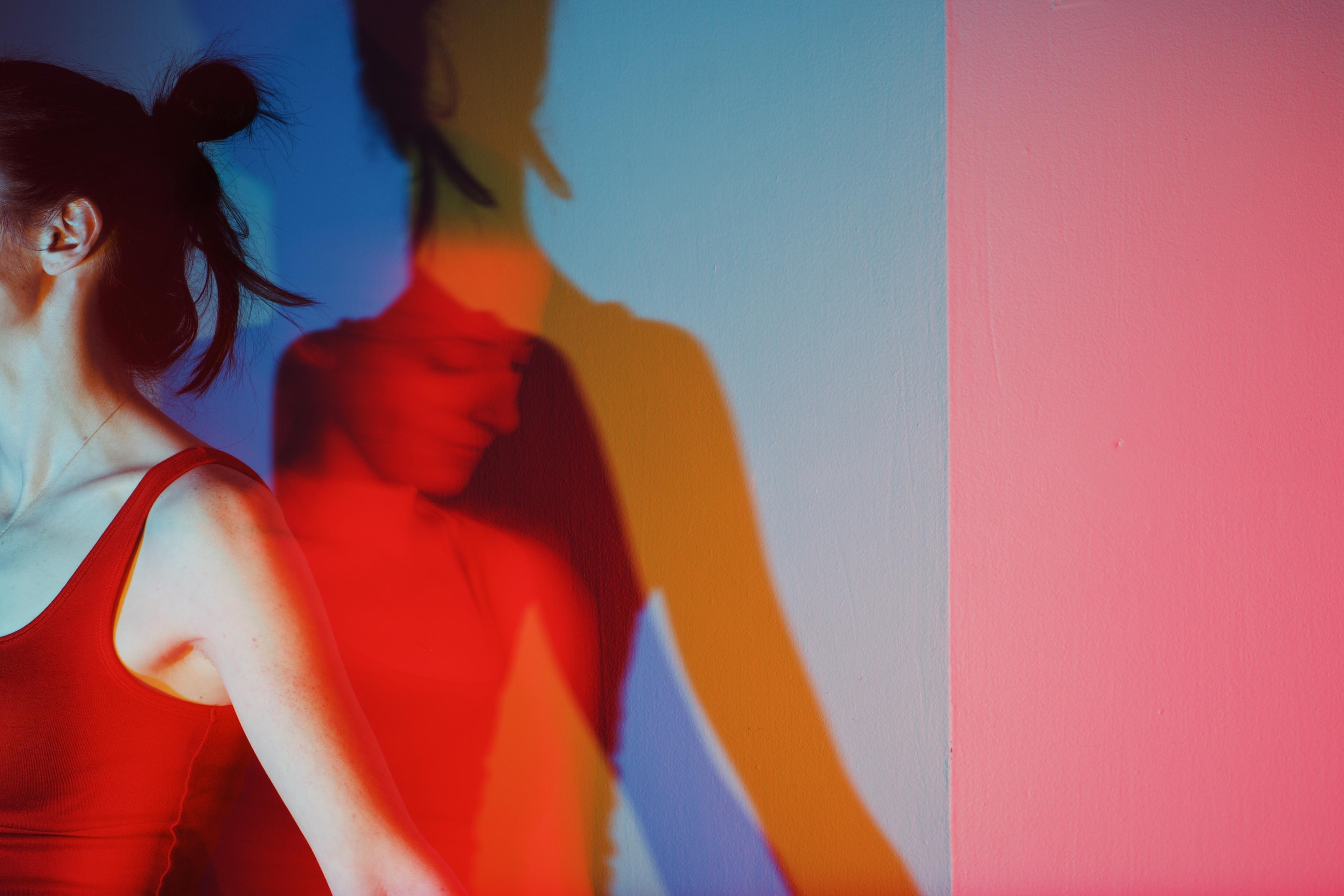 Photo of a woman in a red top with colourful overlapping shadows on a wall displaying red, blue and yellow hues.