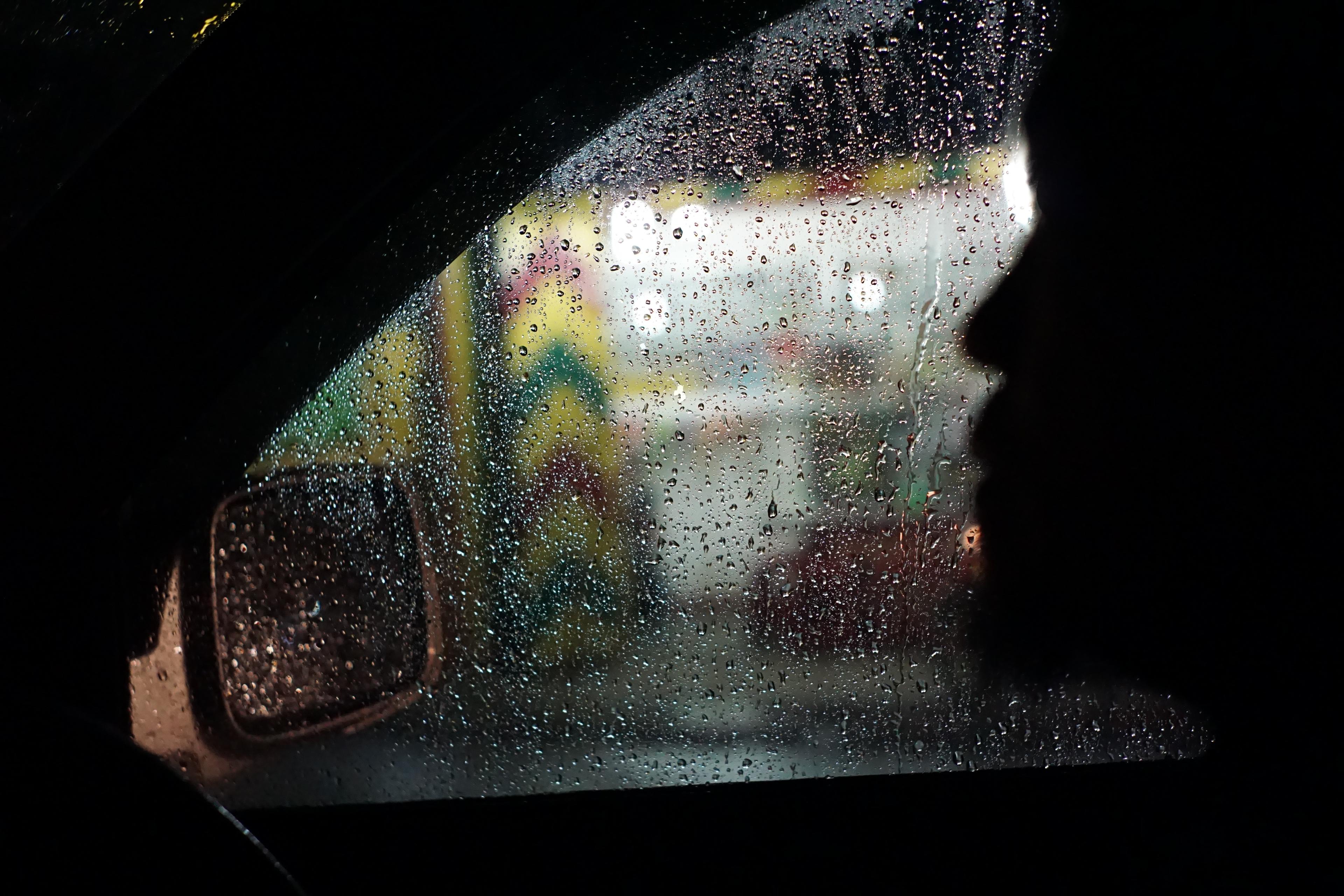 Photo of a rainy car window with a blurred face silhouette inside and colourful lights outside.