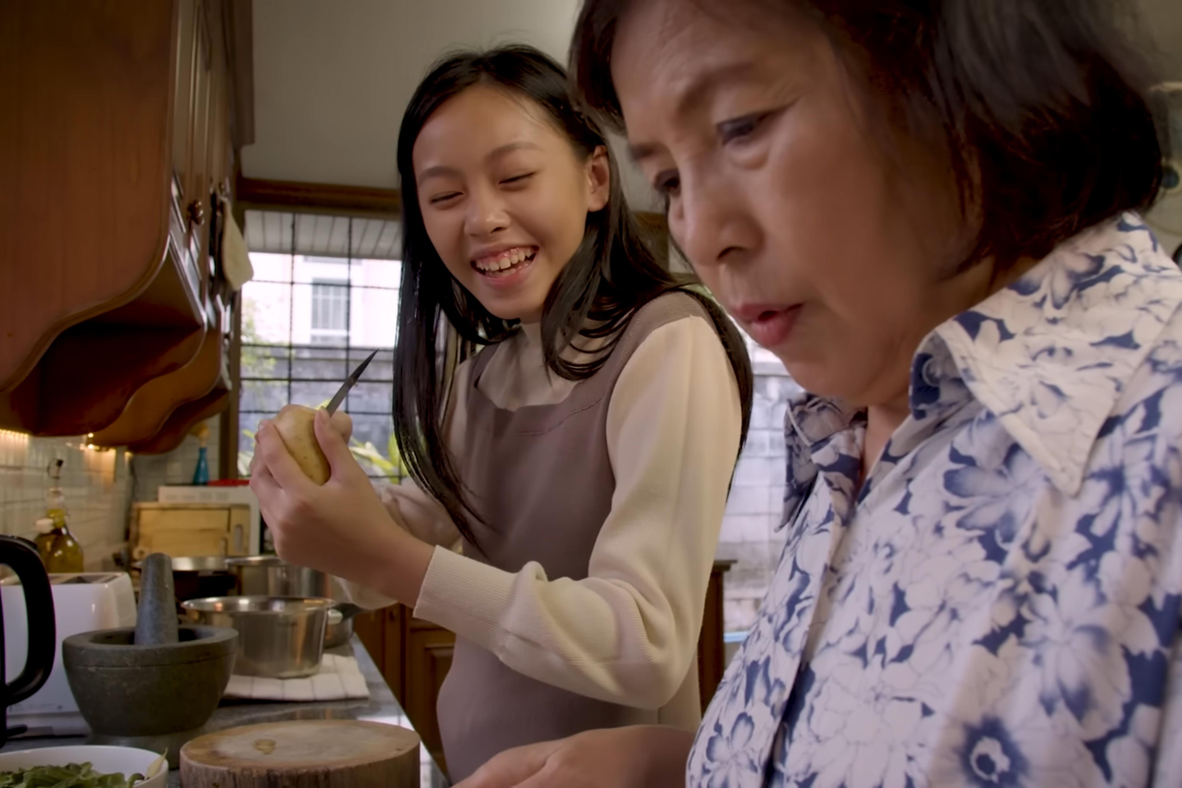 Photo of two women in a kitchen, one peeling a potato while smiling the other focused on cooking, with a mortar in view.