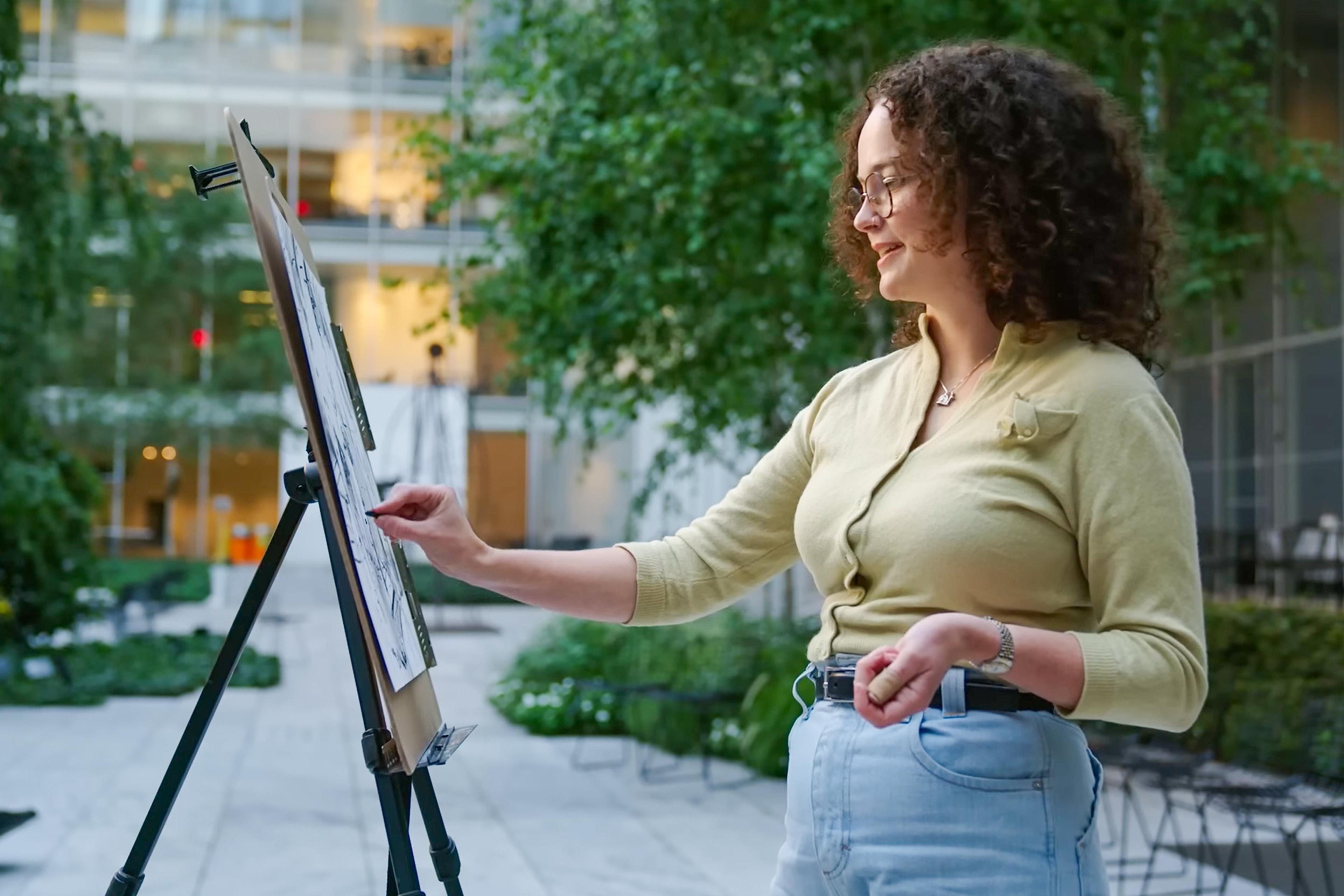 A woman drawing on an easel in a courtyard with greenery and a modern glass building in the background.