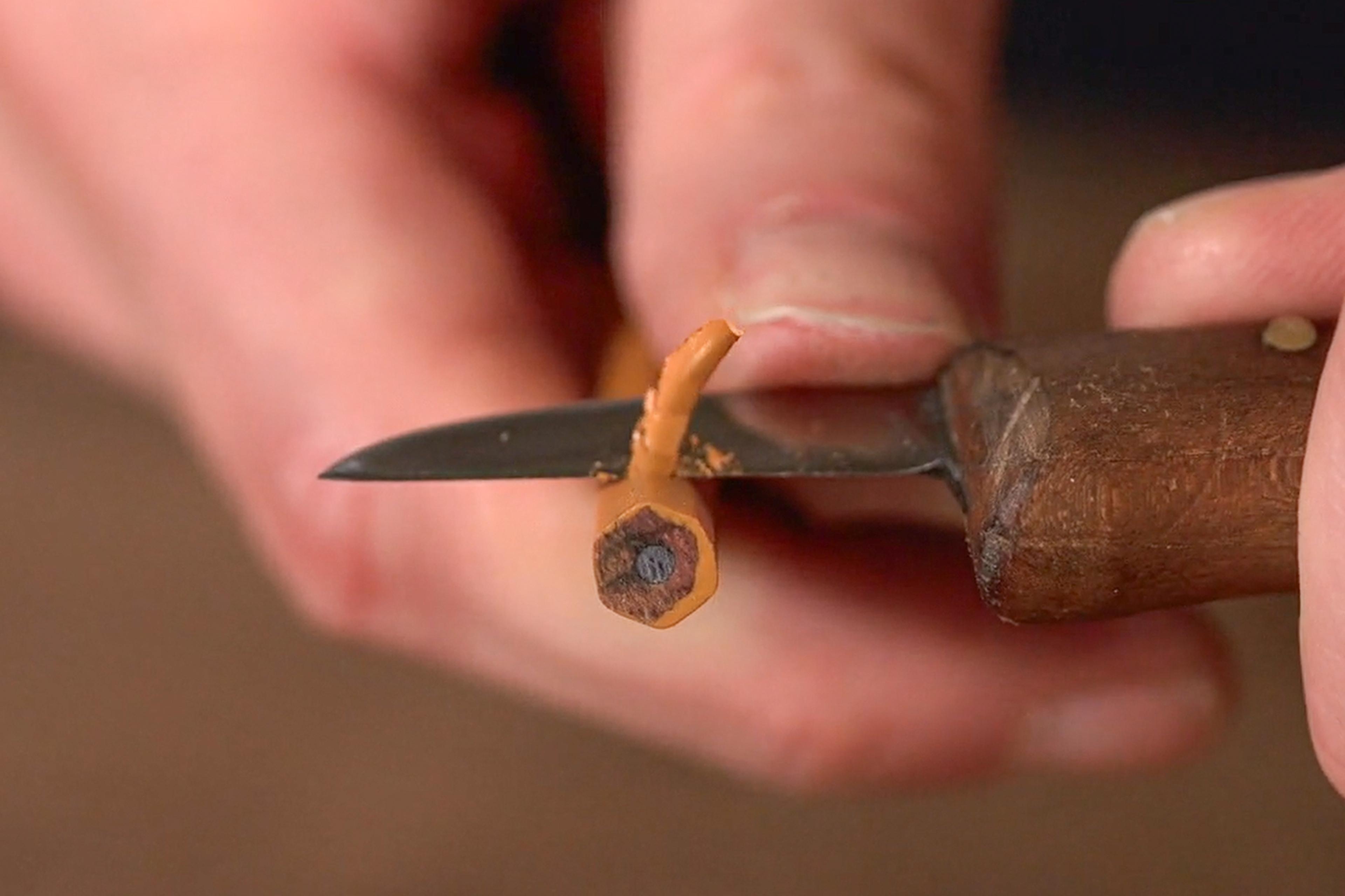 Close-up photo of a hand carving a pencil with a small knife, focusing on the pencil’s tip and wood shavings.