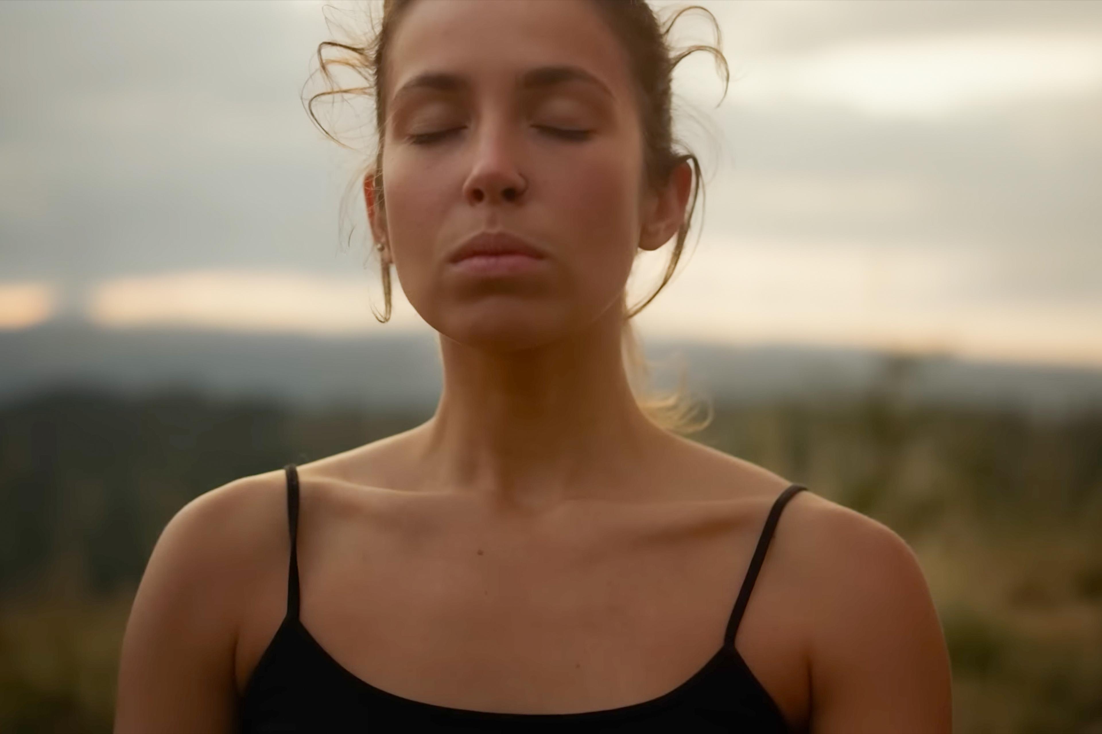 Photo of a serene woman outdoors with closed eyes in a black top, blurred landscape in the background.