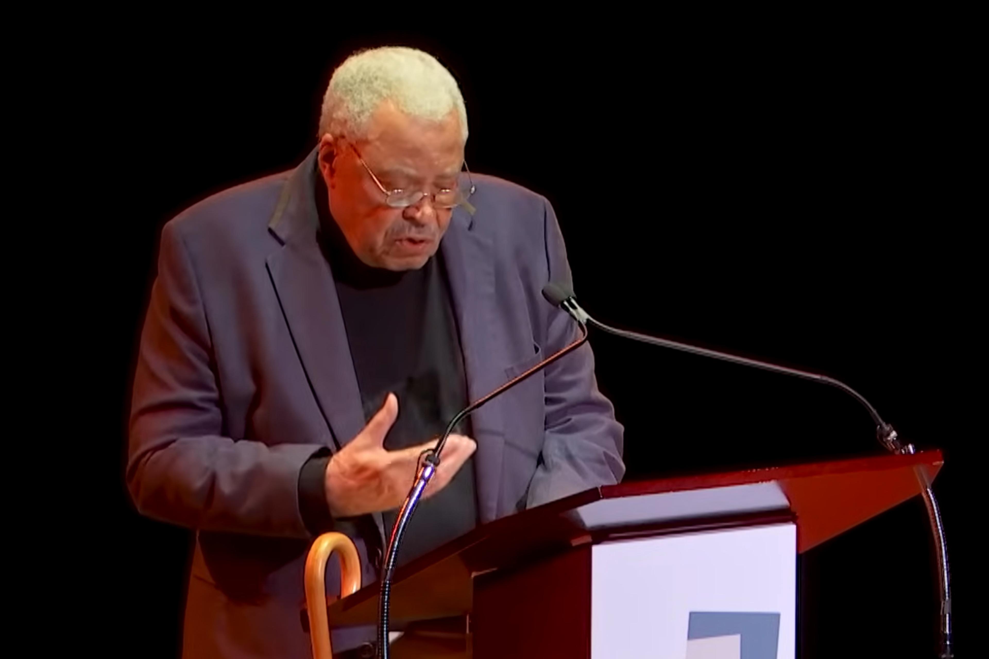 Photo of an elderly man speaking at a podium with a microphone onstage, wearing a dark suit and glasses.