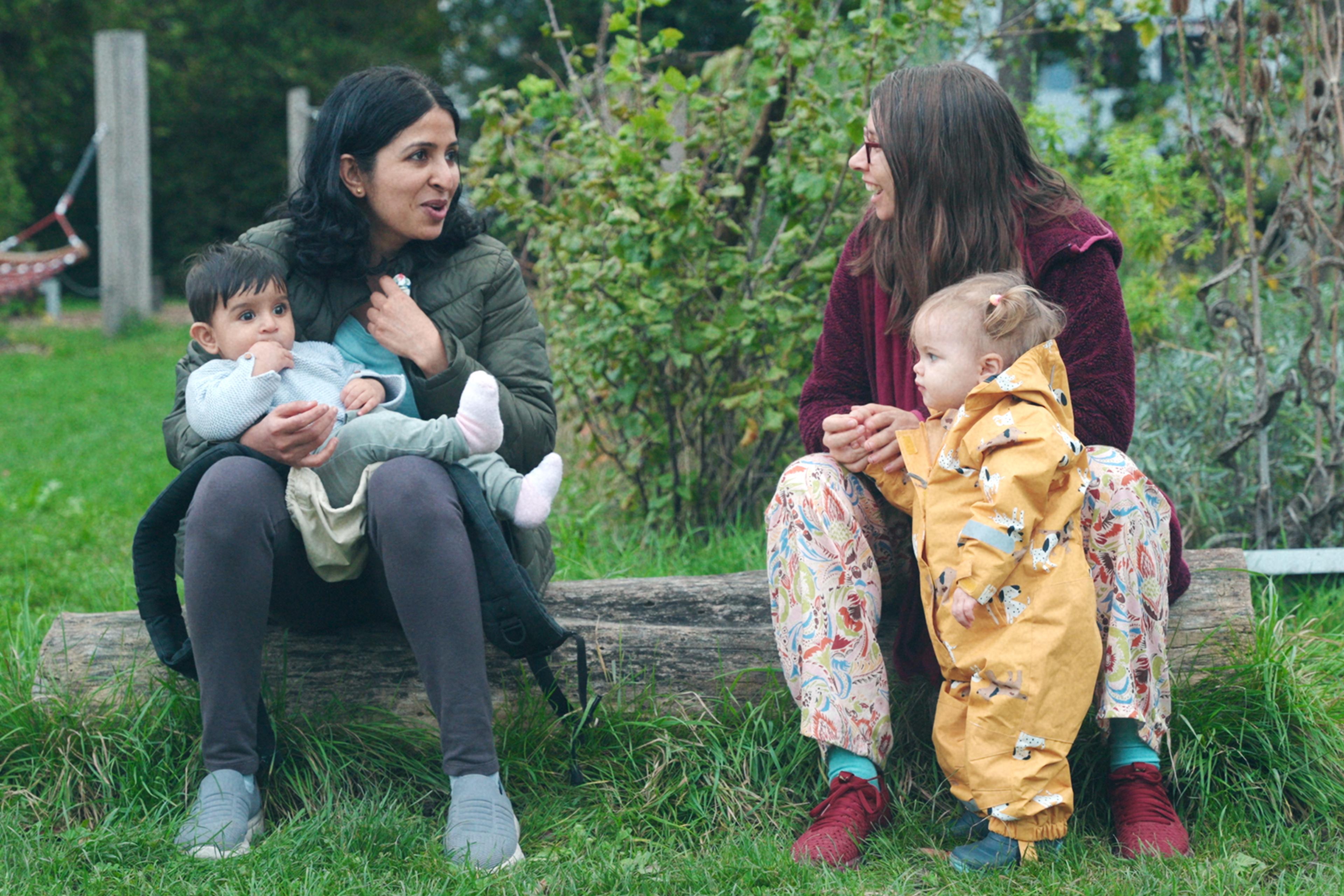 Two women sitting on a log outdoors, each holding a child, with greenery in the background.