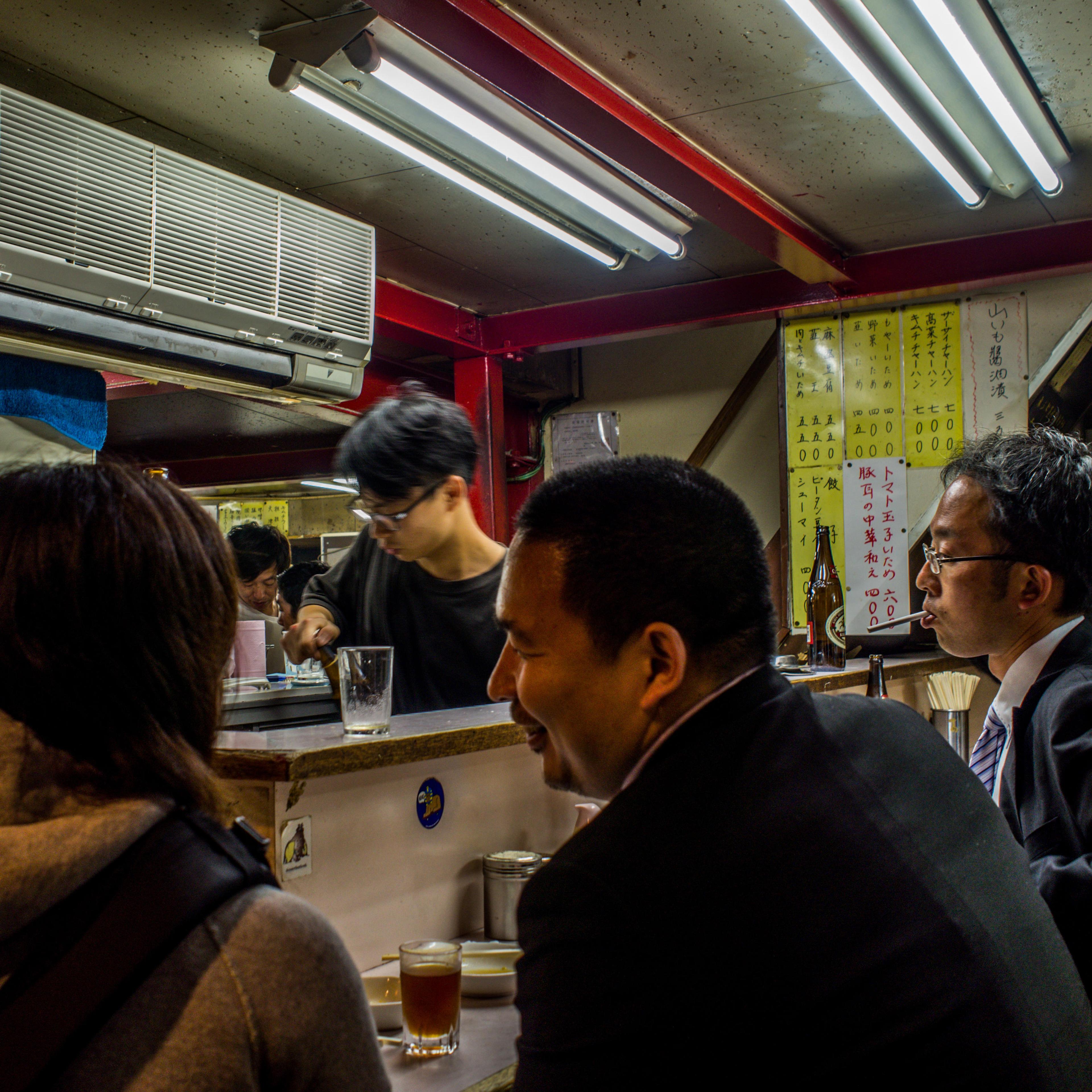 People in a small, well-lit Japanese restaurant or bar, with a menu on the wall and beverages on the counter.