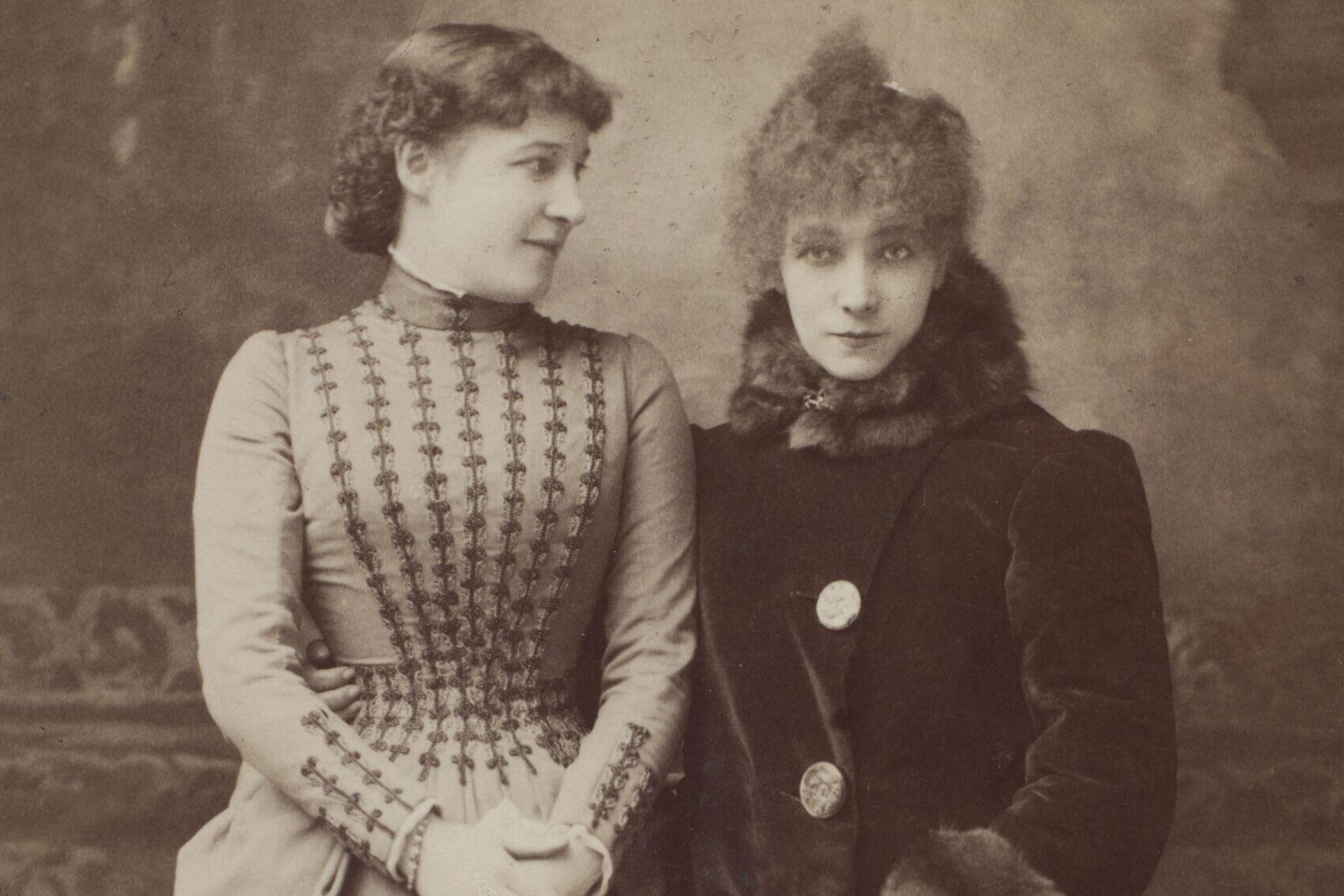 Vintage photo of two women in Victorian attire, one in a patterned dress looks at the other who wears a fur-trimmed coat.