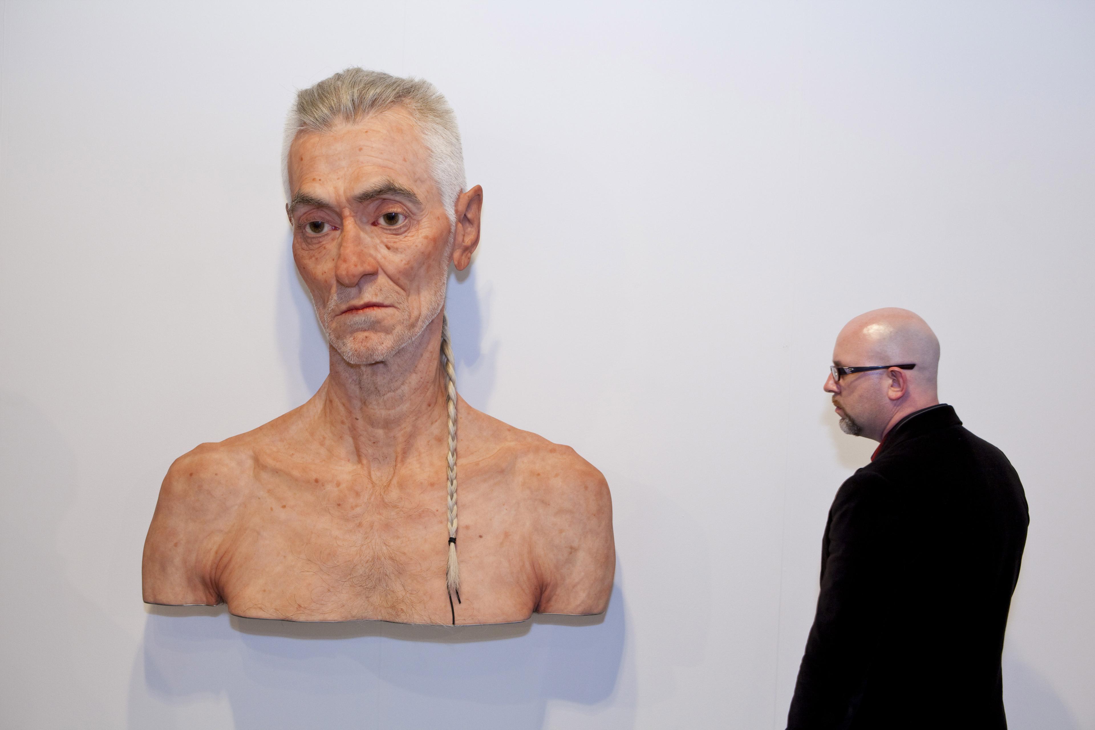A man observing a large, lifelike sculpture of an elderly man with a braid on a white gallery wall.