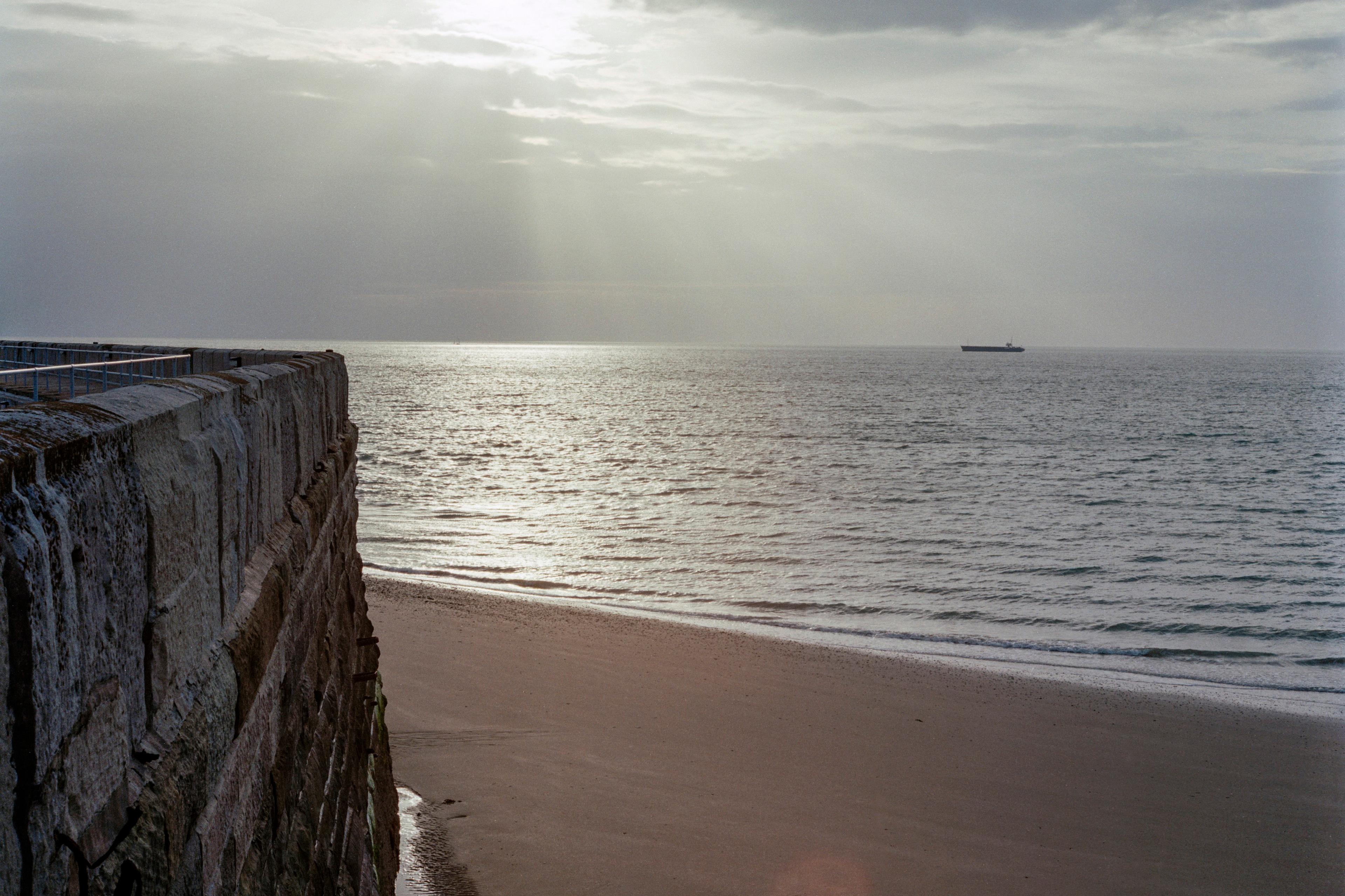 Photo of a stone wall beside a sandy beach with calm sea under a cloudy sky sun peeking through a ship on the horizon.
