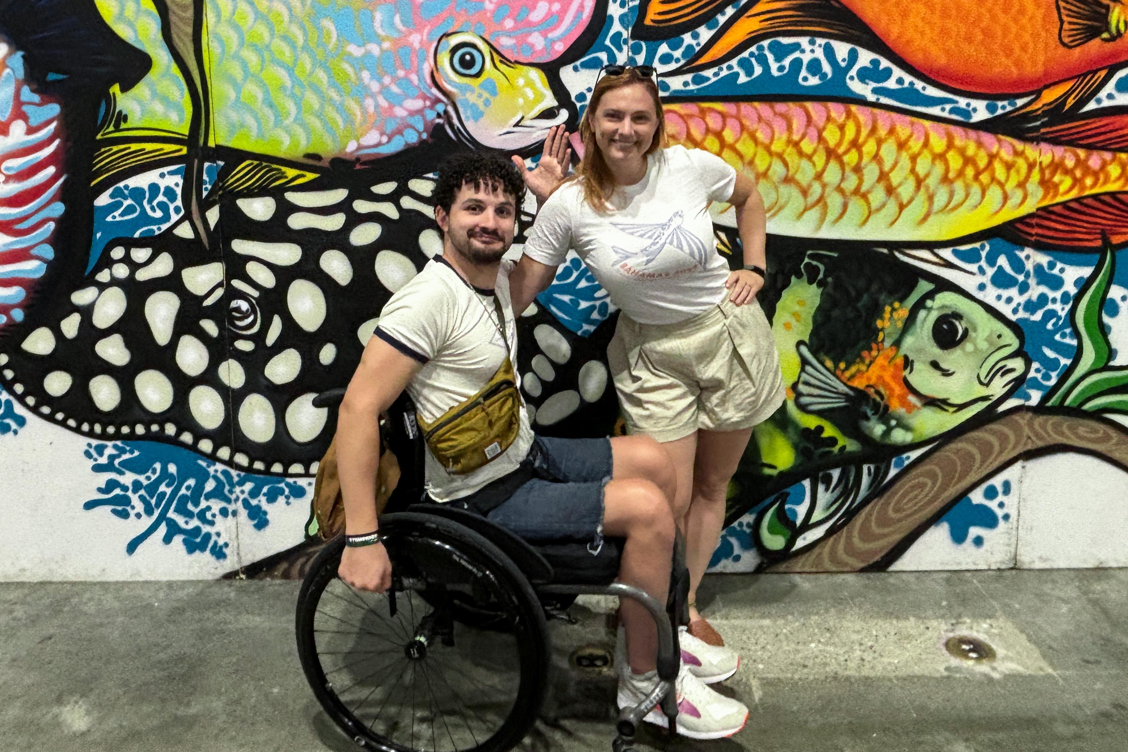 Two people smiling in front of colourful fish graffiti, the man is sitting in a wheelchair the woman is standing beside him.