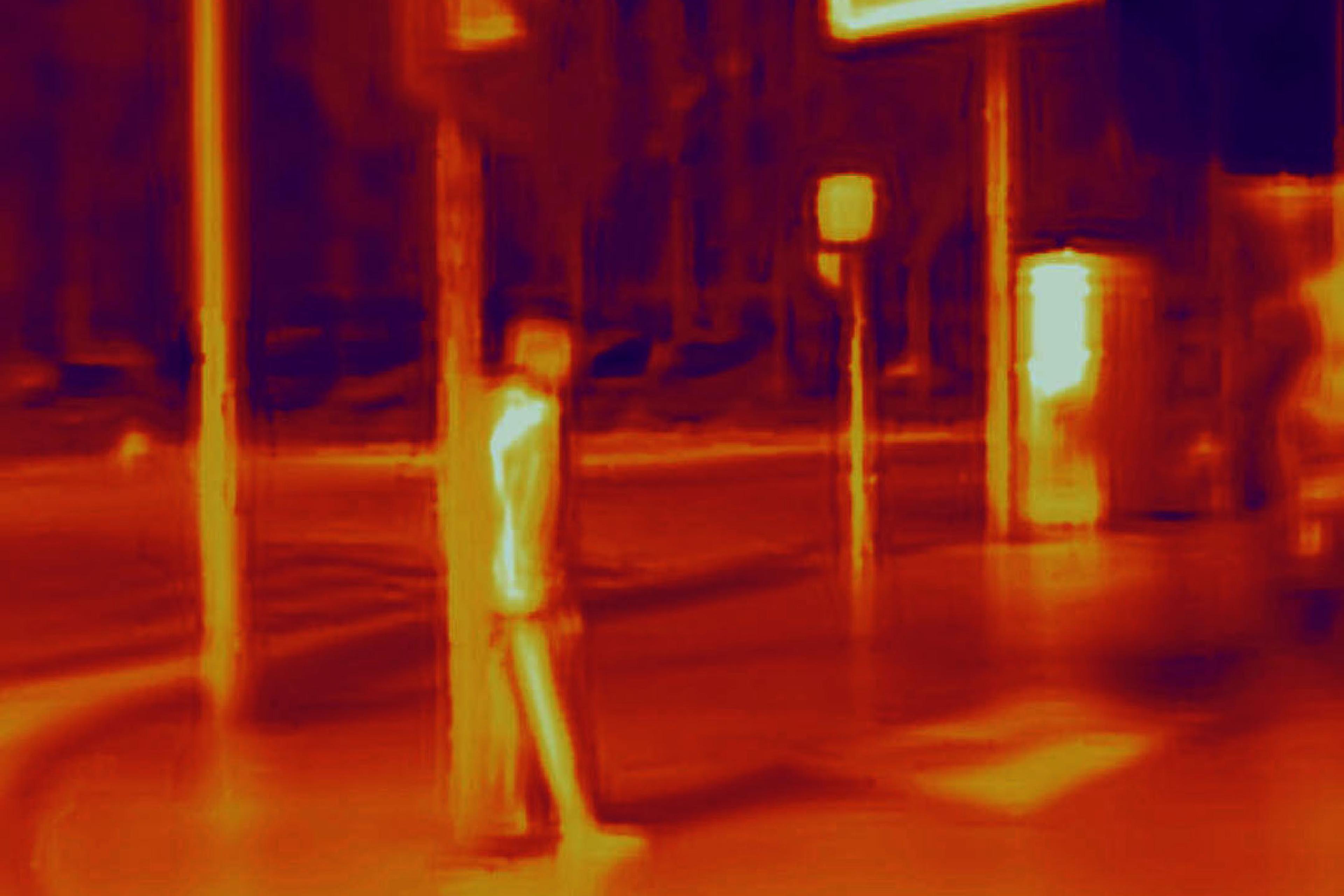 Thermal image of a street scene with a person standing by a pole and bright signage in the background.