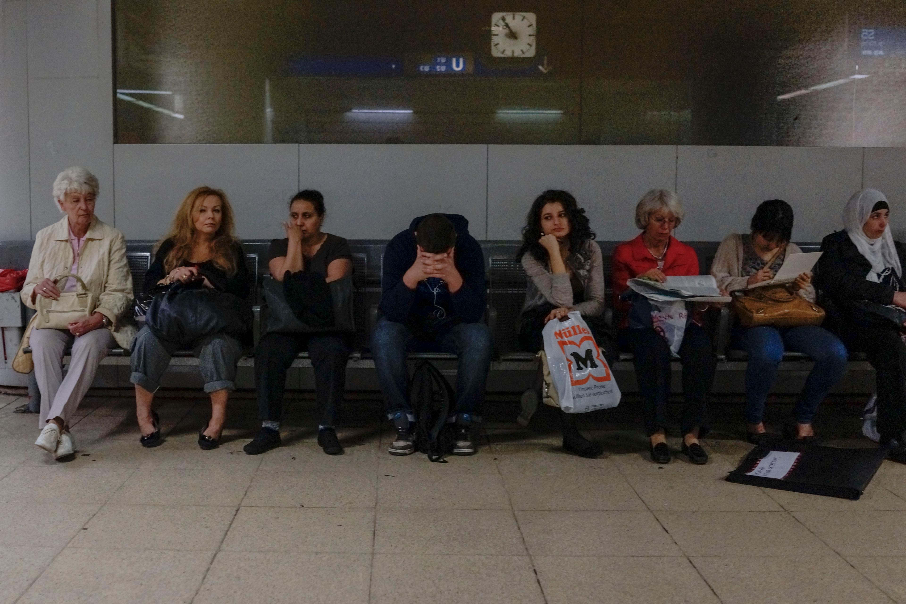 Photo of seven people sitting on a bench in a train station waiting area, with a clock on the wall behind them.