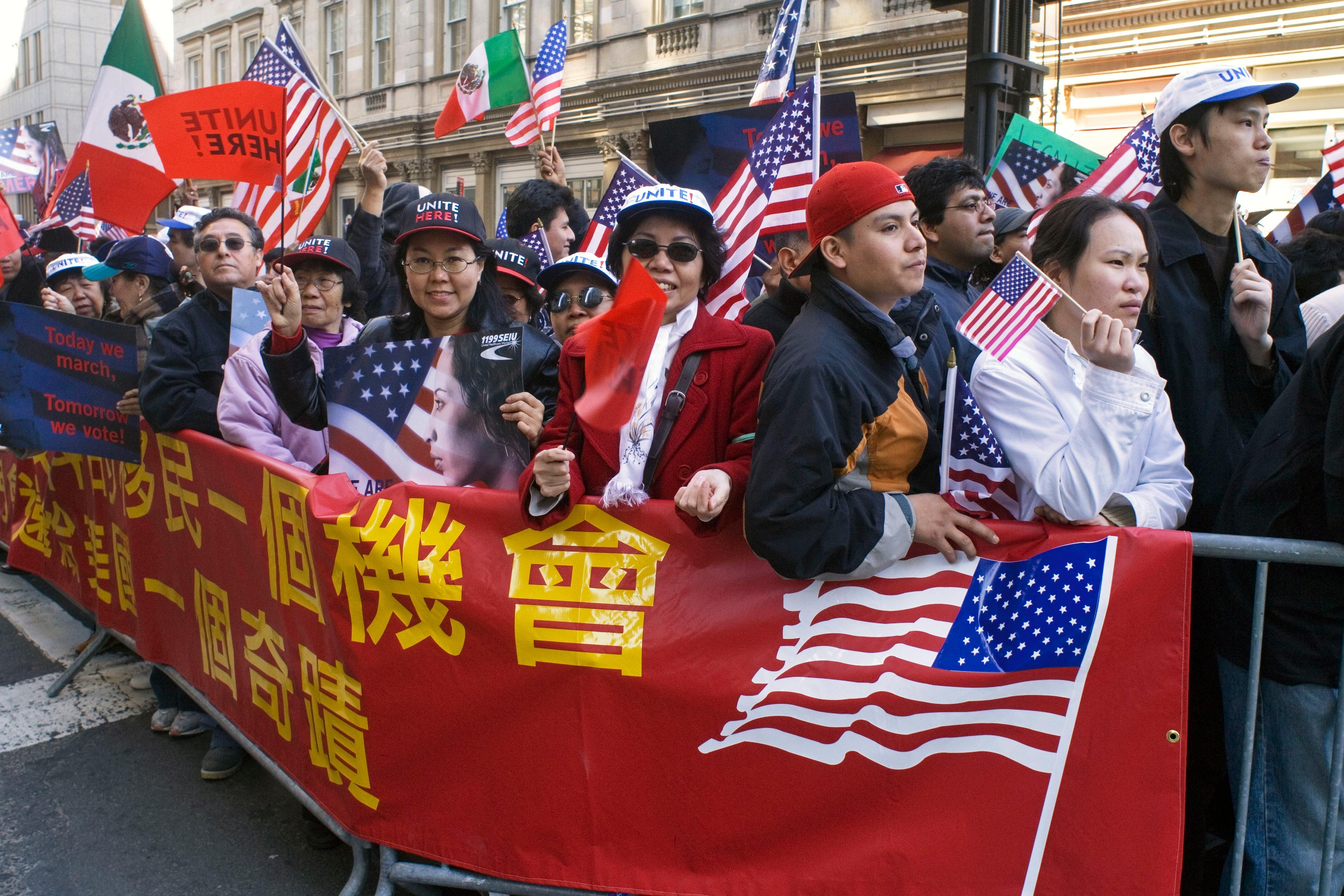 Photo of a diverse crowd holding American flags and banners during a street protest, with buildings in the background.