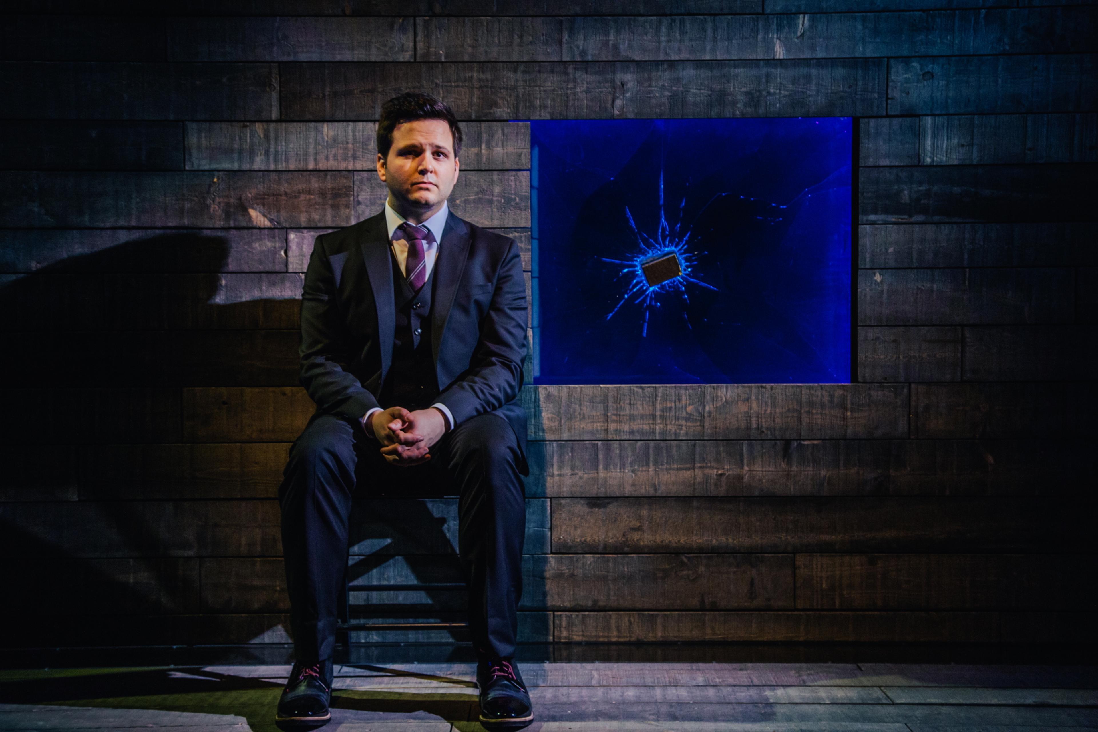 Photo of a man in a suit sitting in a dimly lit room with a shattered blue screen on a wooden wall behind him.