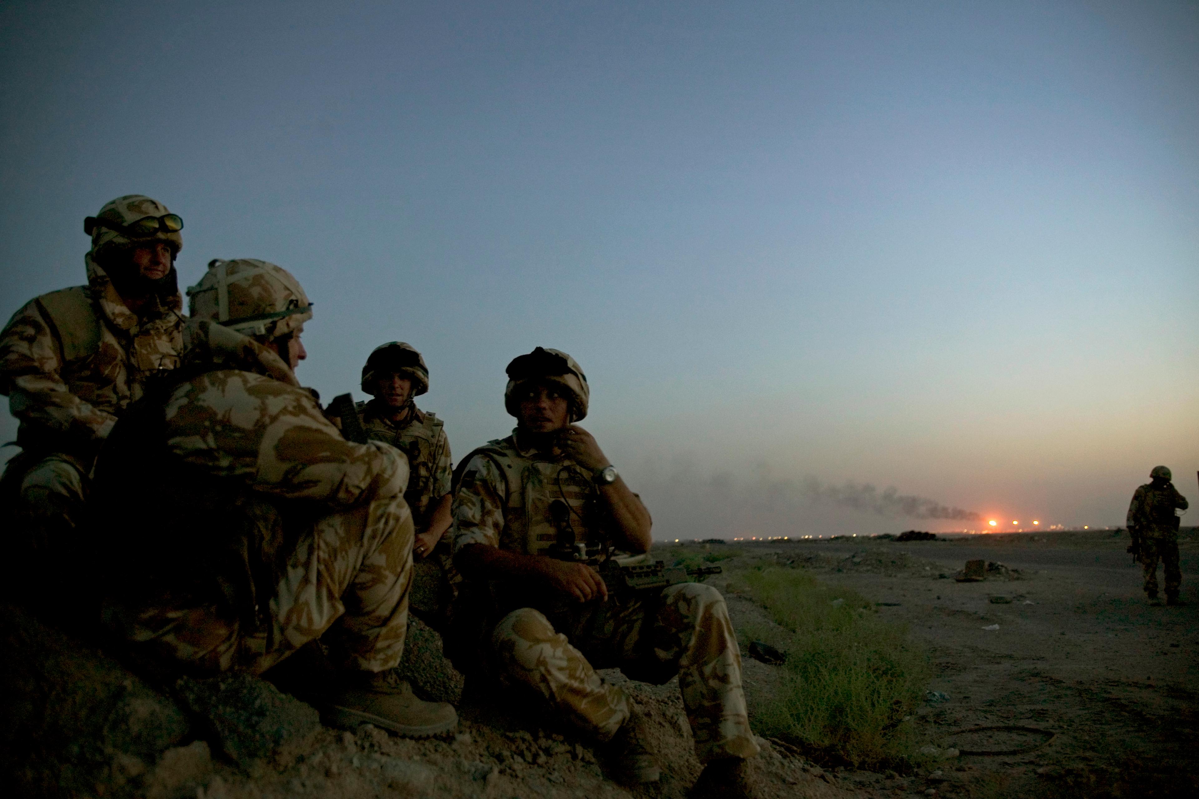 Photo of soldiers in camouflage sitting on a rocky terrain at dusk with a smoky horizon and distant lights in the background.