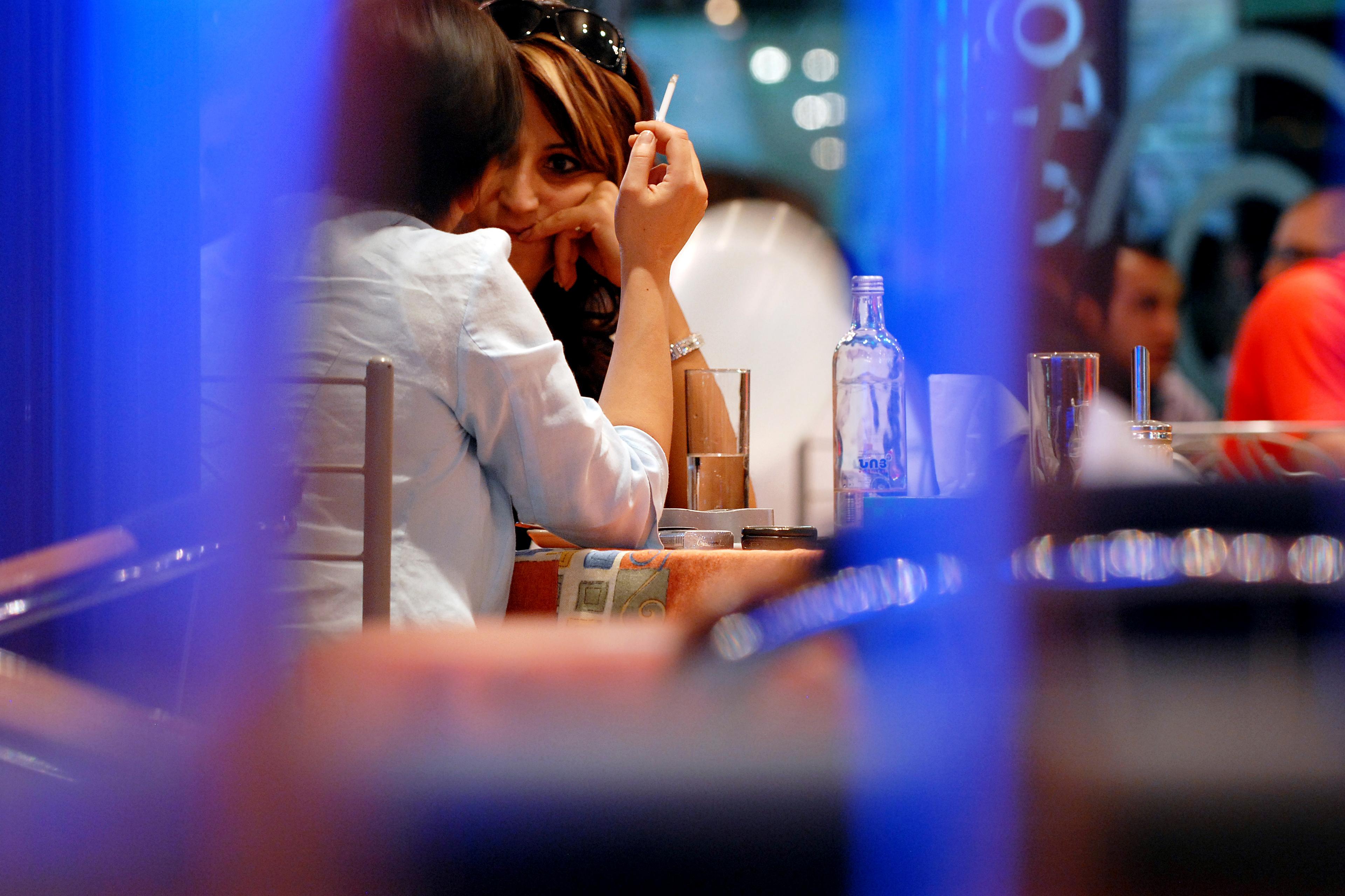 Two people at a cafe table, one holding a cigarette. Blurred foreground and background create a candid atmosphere.