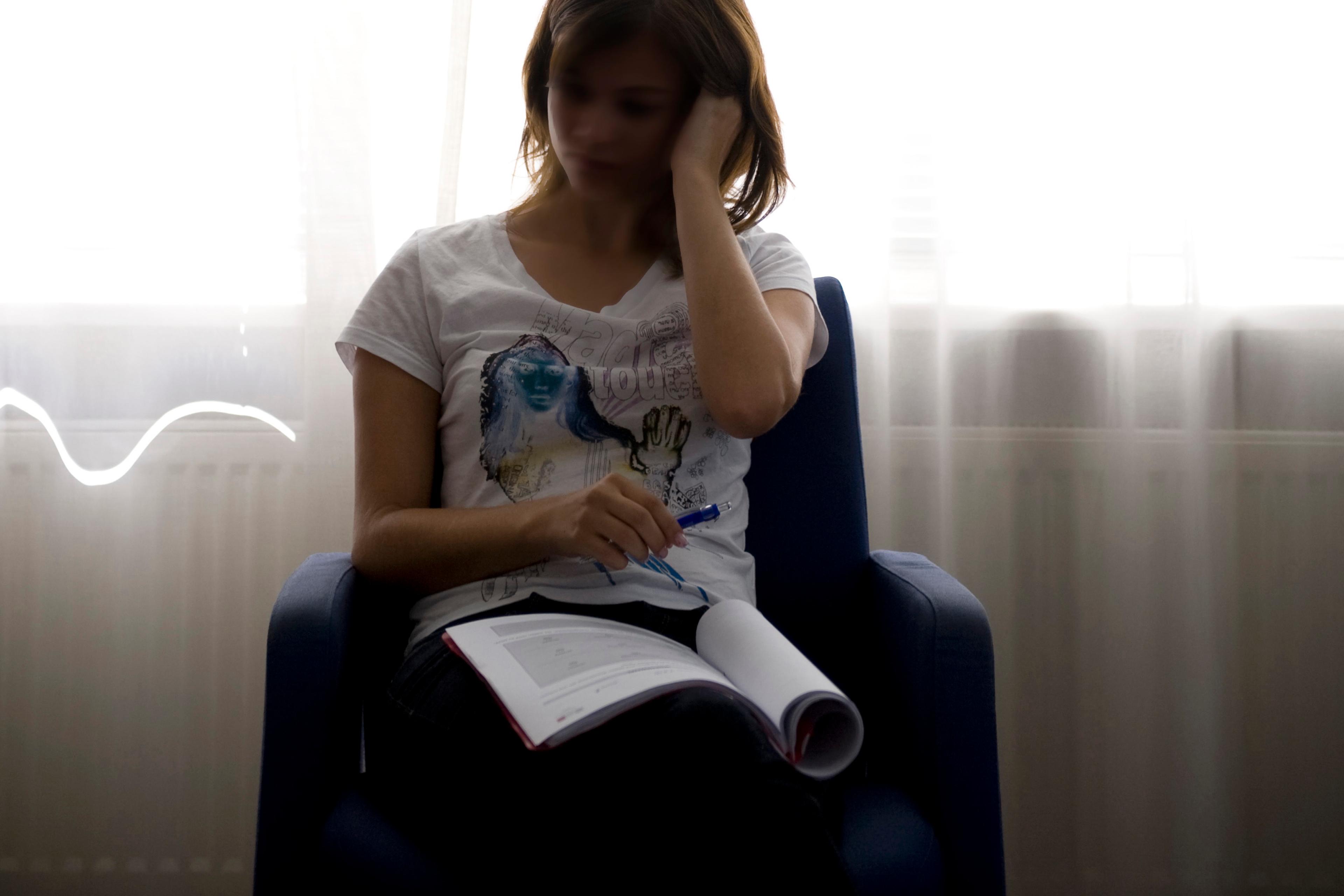 Photo of a woman sitting by a bright window reading a book in a relaxed pose.