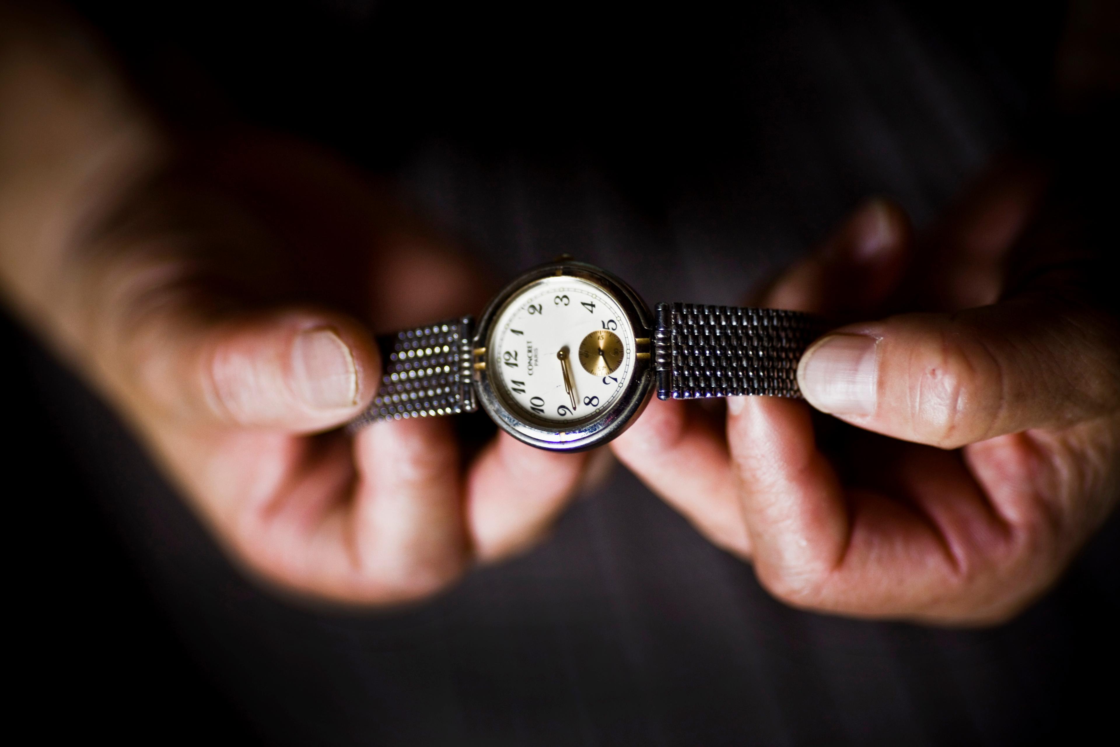 Close-up photo of hands holding a silver wristwatch with a white dial and black numerals on a black background.
